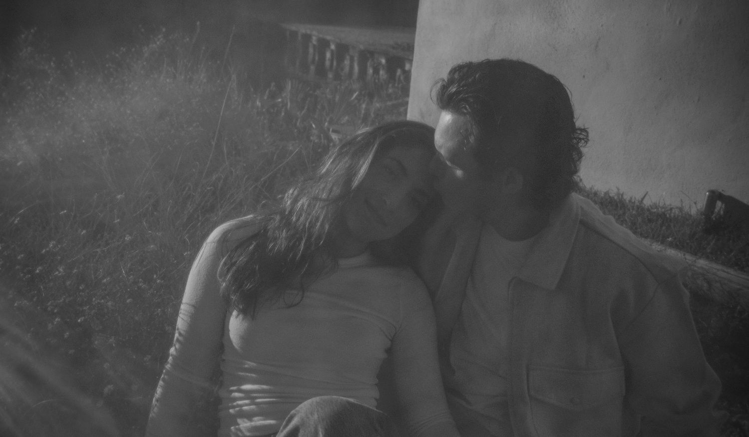 Black and white photo of a couple sitting close on the grass, her head resting on his shoulder.
