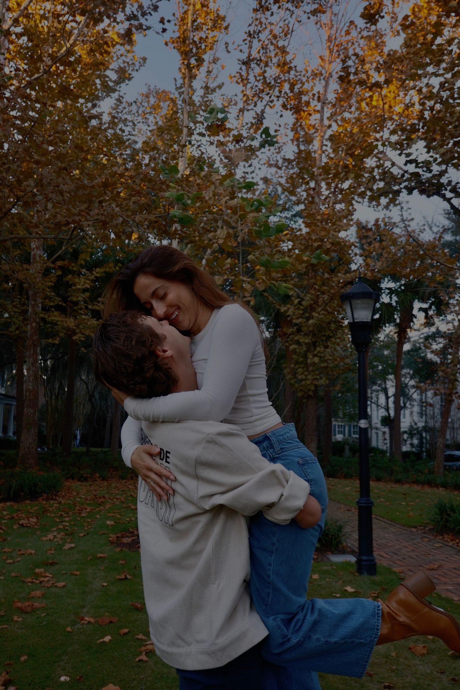 A man lifts a laughing woman in an embrace under autumn trees beside a lamp post on a leafy park path.