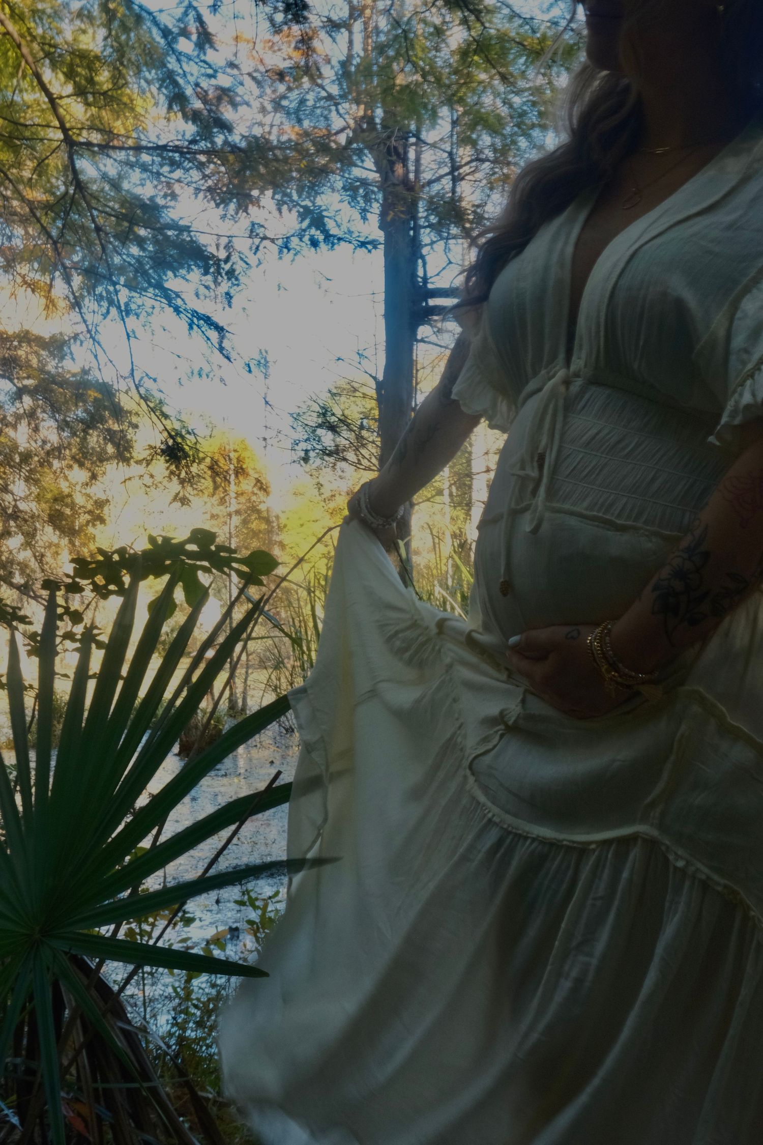 A pregnant woman in a flowing white boho dress stands in a lush forest by a palmetto plant, gently holding her skirt.