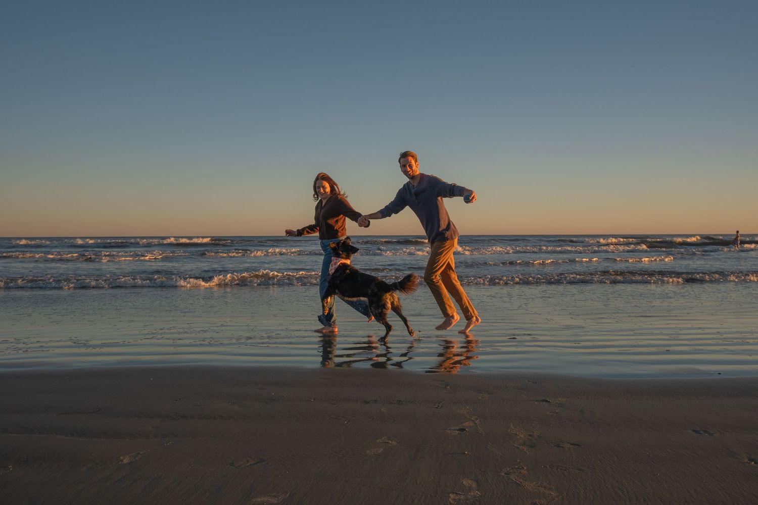Couple holding hands and playing with their dog in shallow beach waves at sunset under a clear gradient sky.