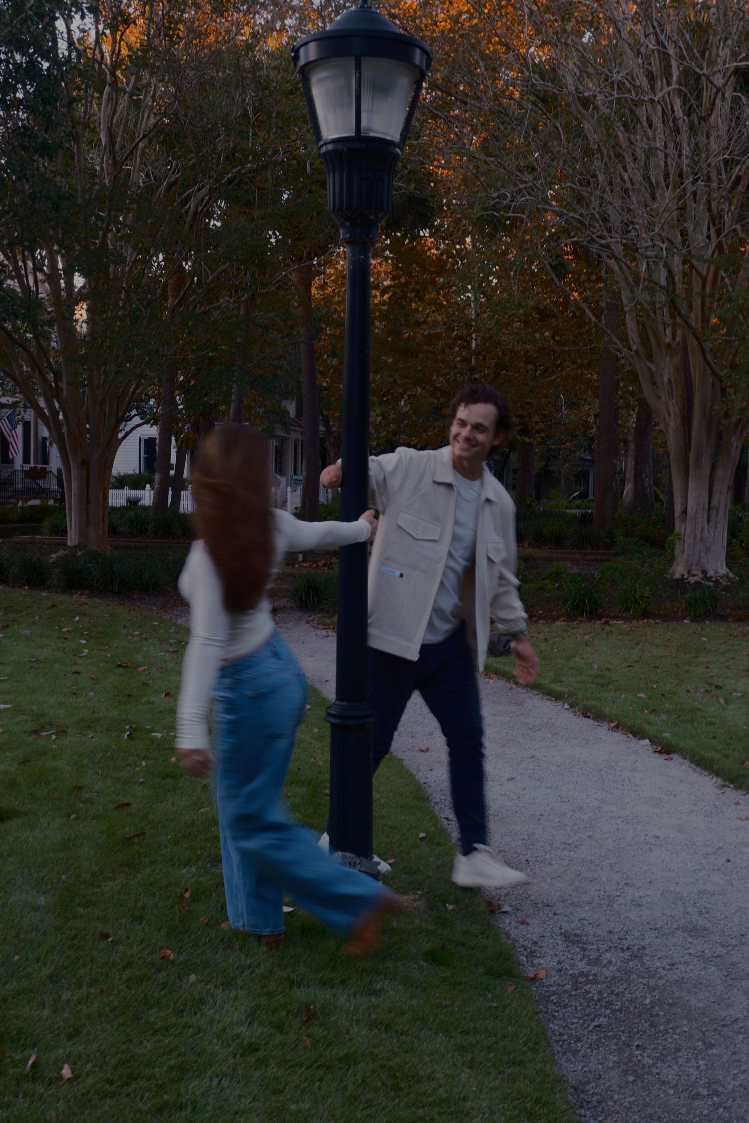 Smiling man in a cream jacket reaches toward a woman walking around a lamp post on a park path in autumn.