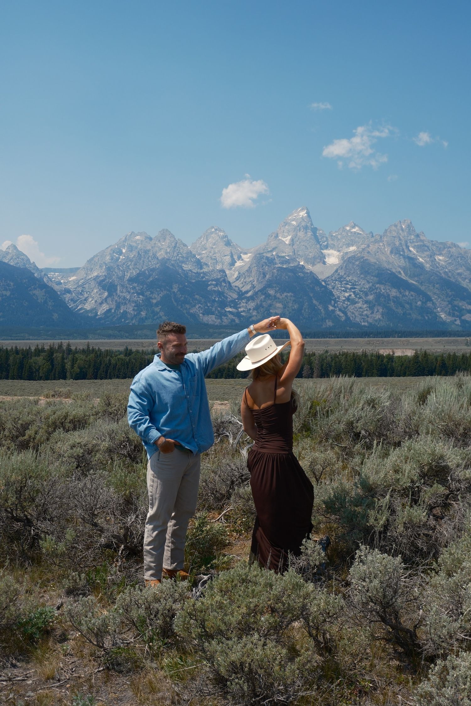Man in a blue shirt twirls a woman in a brown dress and cowboy hat in a sagebrush field with snowy mountains behind them.