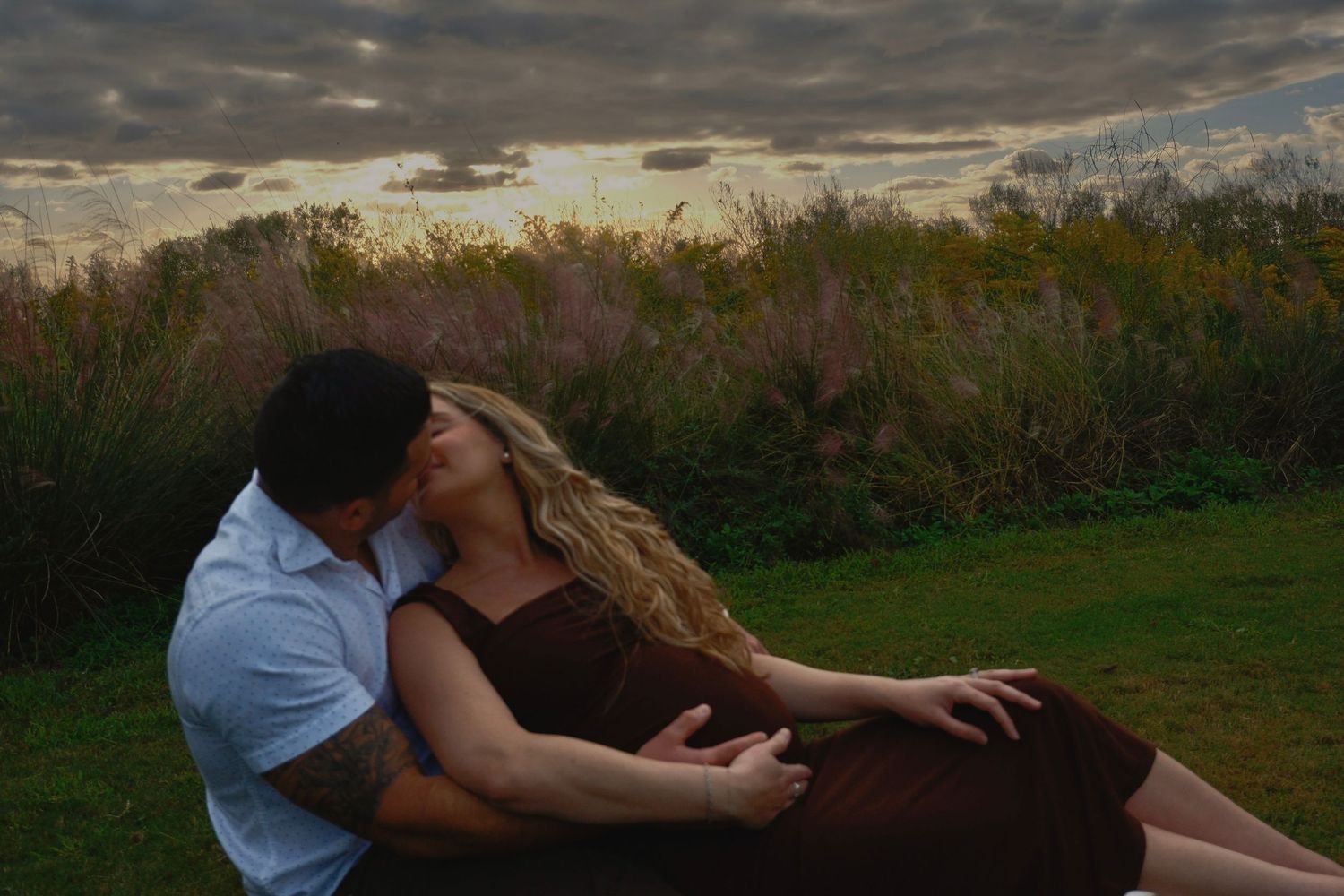 Couple kissing on the grass at sunset, the woman in a dark dress with long blonde hair, wild field behind them.