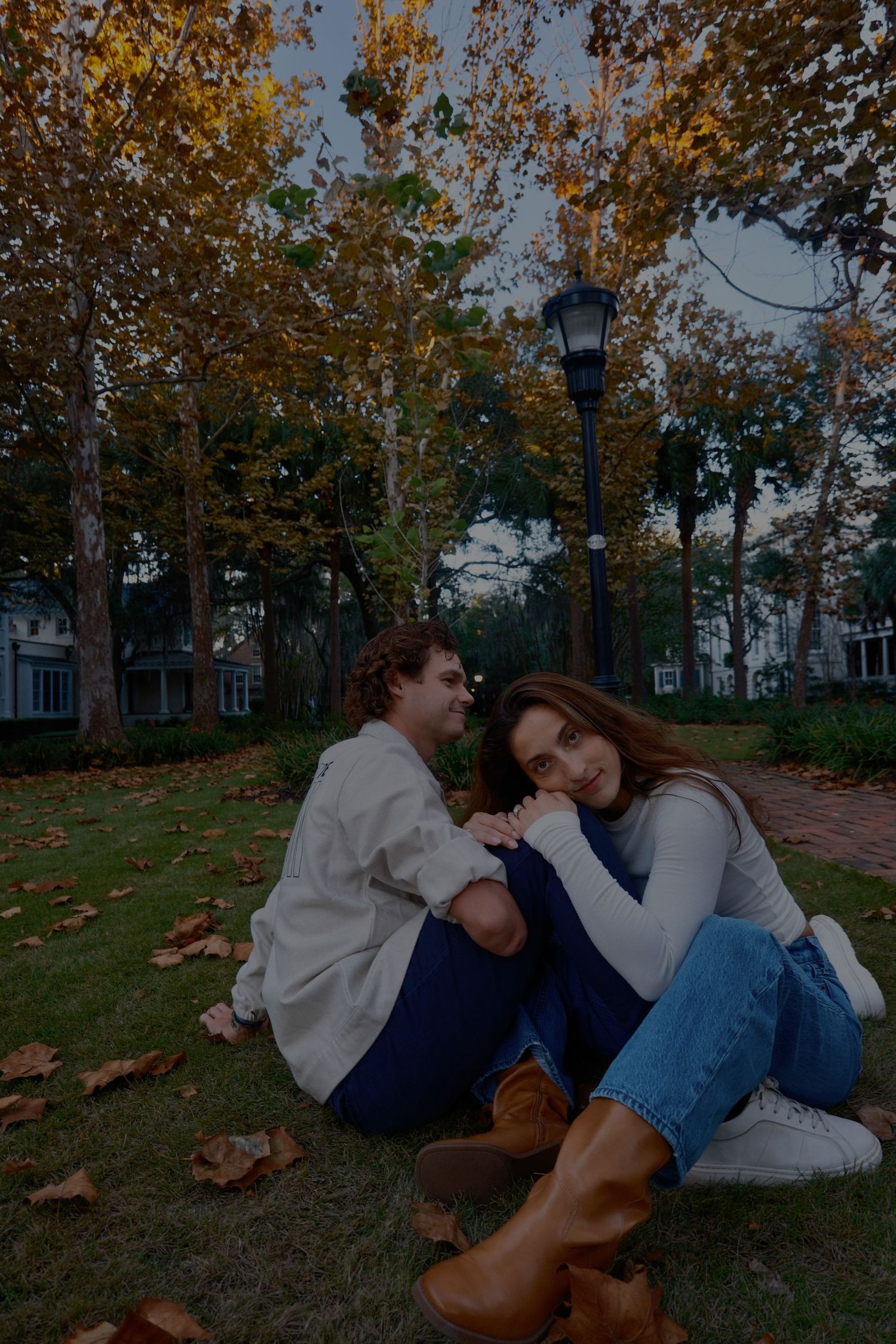 Couple sits together on a leaf-covered lawn under autumn trees, smiling beside a vintage street lamp.