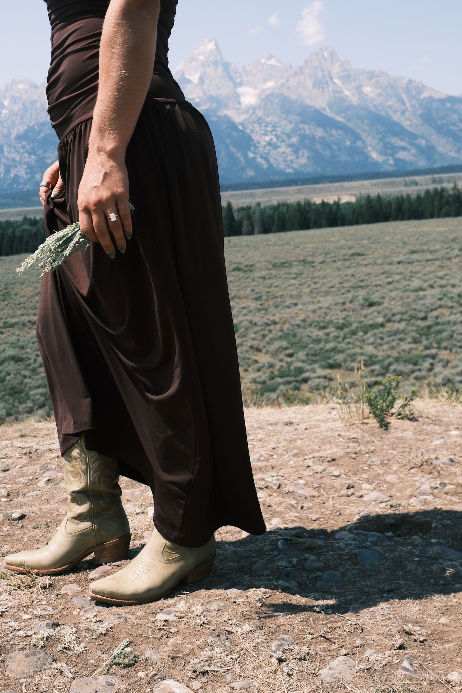 A woman in a long brown skirt and cream cowboy boots holds dried lavender while standing before the snowy Teton mountain range.