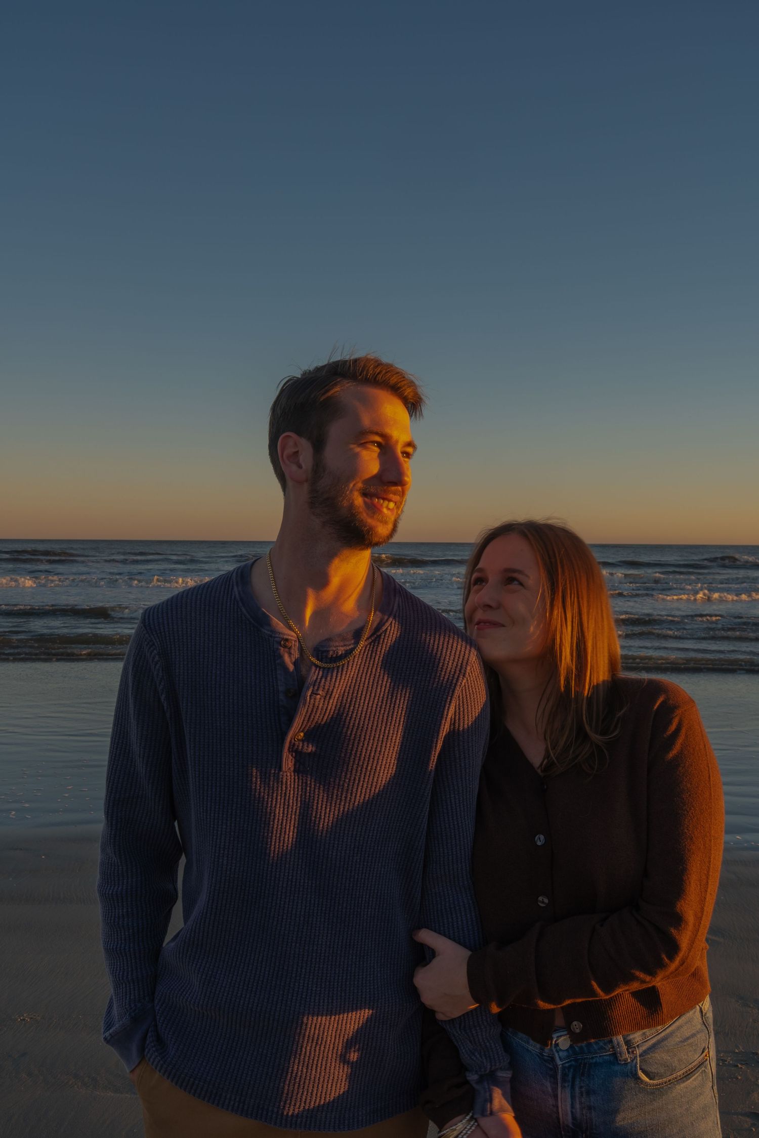 Smiling couple embraces on the beach at golden hour with the ocean and a clear sunset sky behind them.