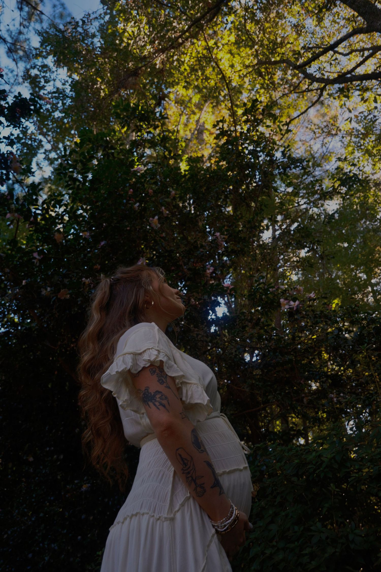 Pregnant woman with tattoos and wavy hair in a white ruffled dress looking up at tall sunlit trees.