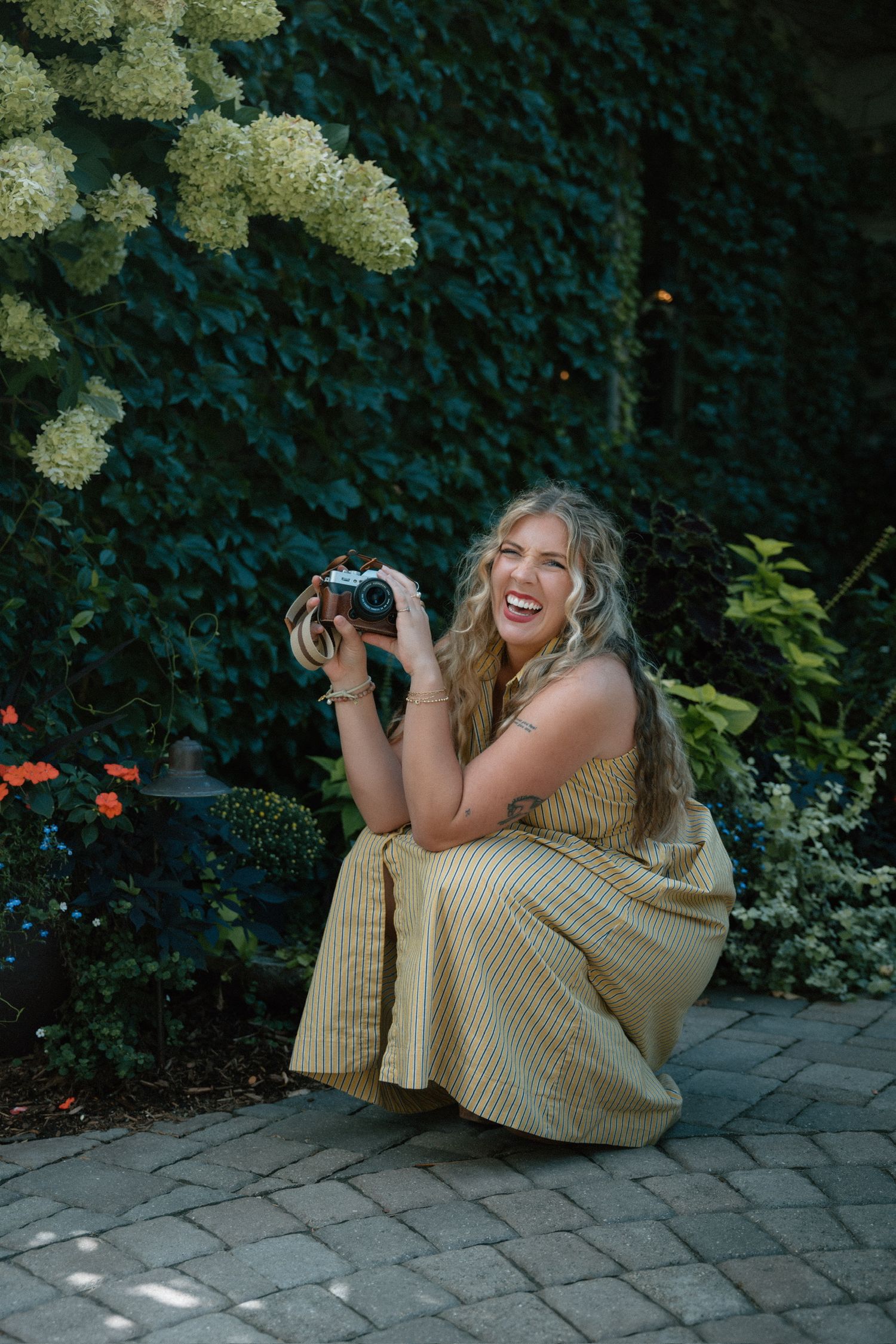 Laughing woman in a yellow striped dress crouches on a brick patio holding a film camera near blooming white hydrangeas.