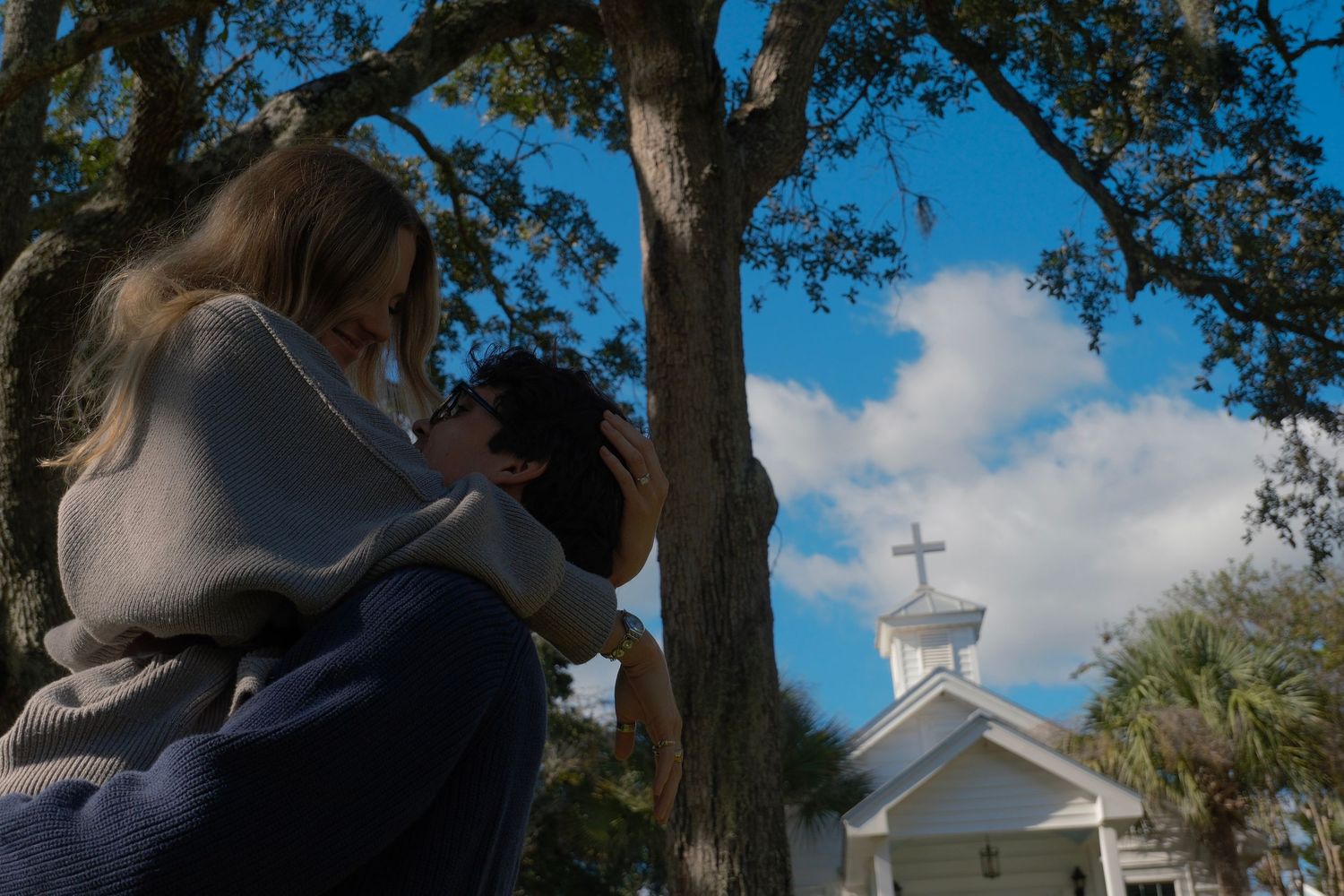 Woman in a gray sweater holds a child up beneath a large oak tree with a small white chapel steeple in the background.