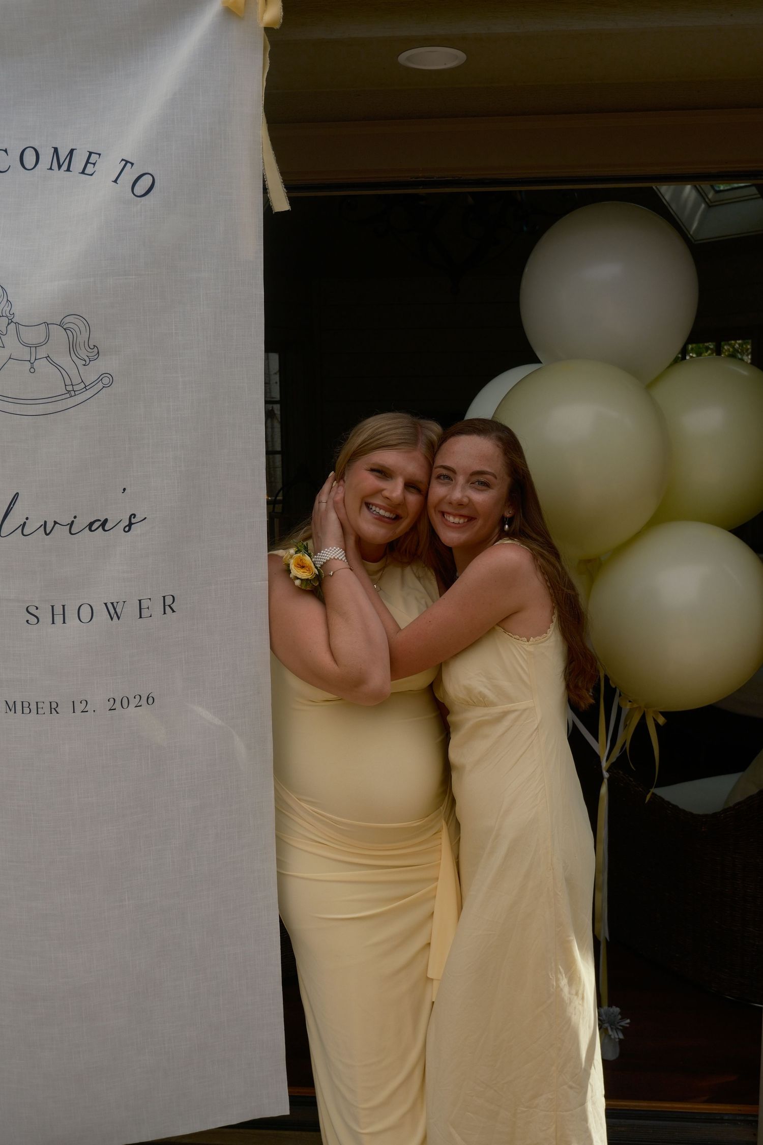Two women in yellow dresses embrace and smile beside a baby shower banner and a cluster of cream and sage balloons.