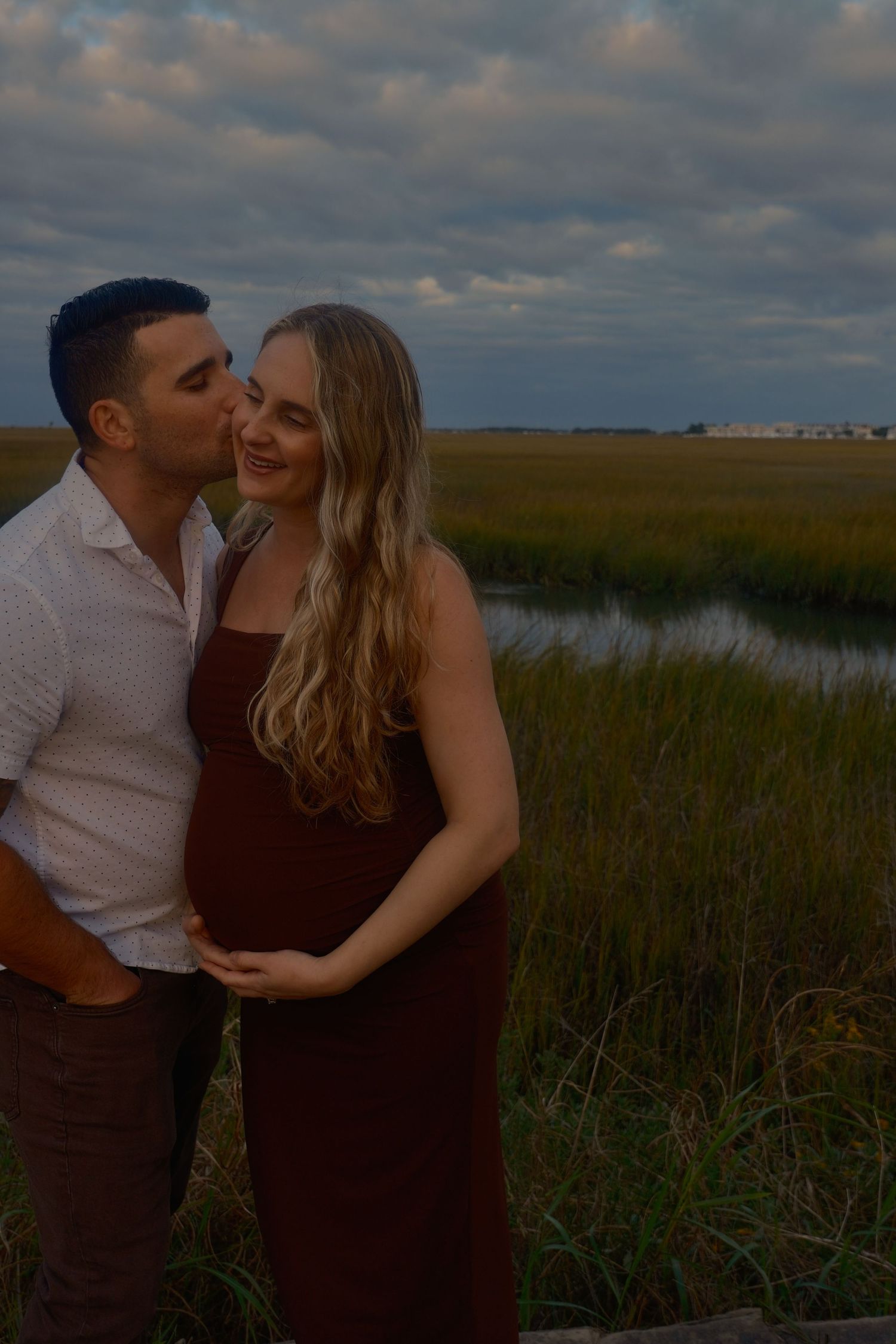 Expectant couple shares a kiss on a marsh boardwalk at sunset, the woman cradling her baby bump in a burgundy dress.