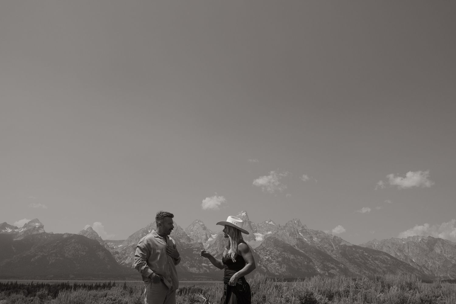 Black and white image of a couple in conversation in an open meadow, with a dramatic mountain range along the horizon.