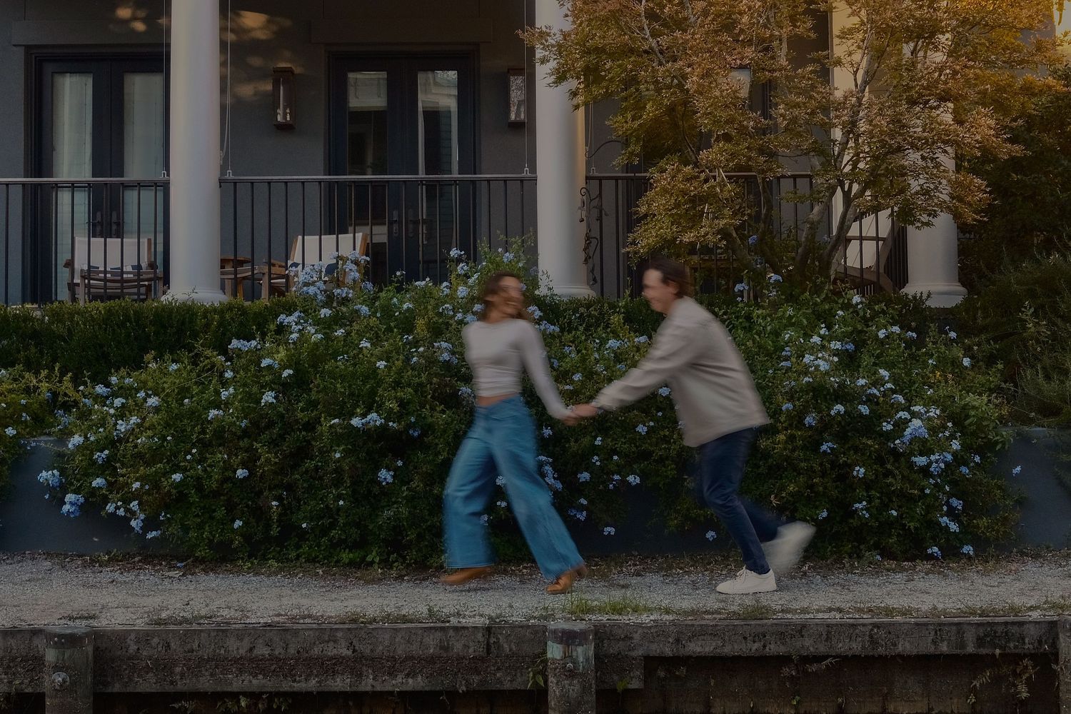 Couple laughing and holding hands while walking past blooming blue hydrangeas in front of a porch.