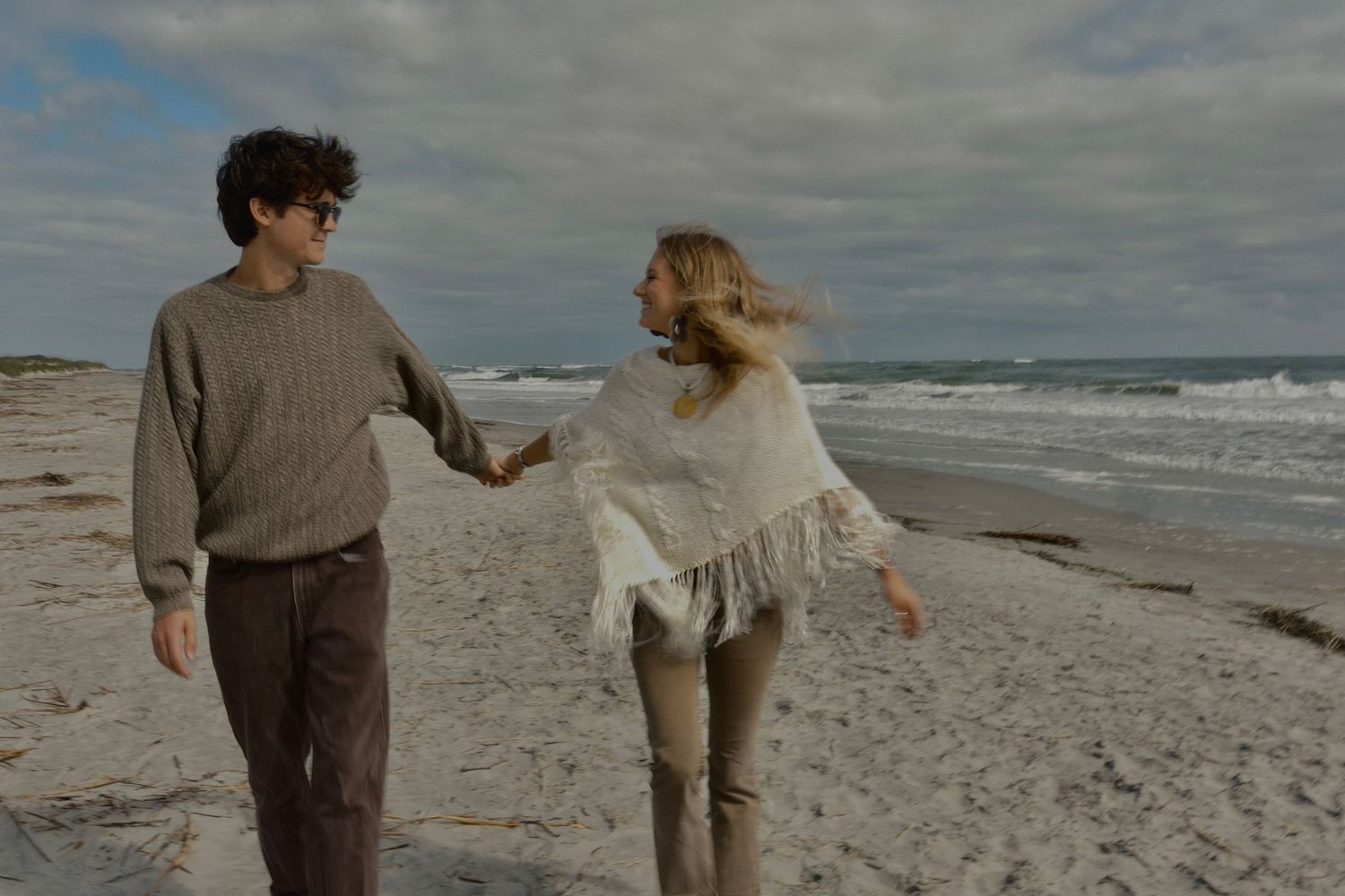 Couple in sweaters walking hand in hand along a beach with waves rolling in under a cloudy sky.