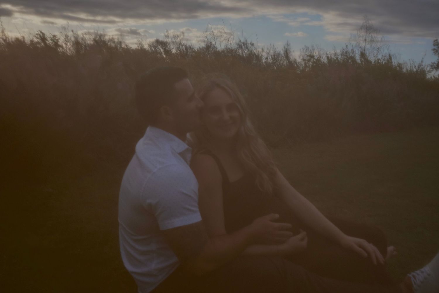 Couple sits close together in a grassy field at dusk, smiling and embracing in a moody, softly lit portrait.