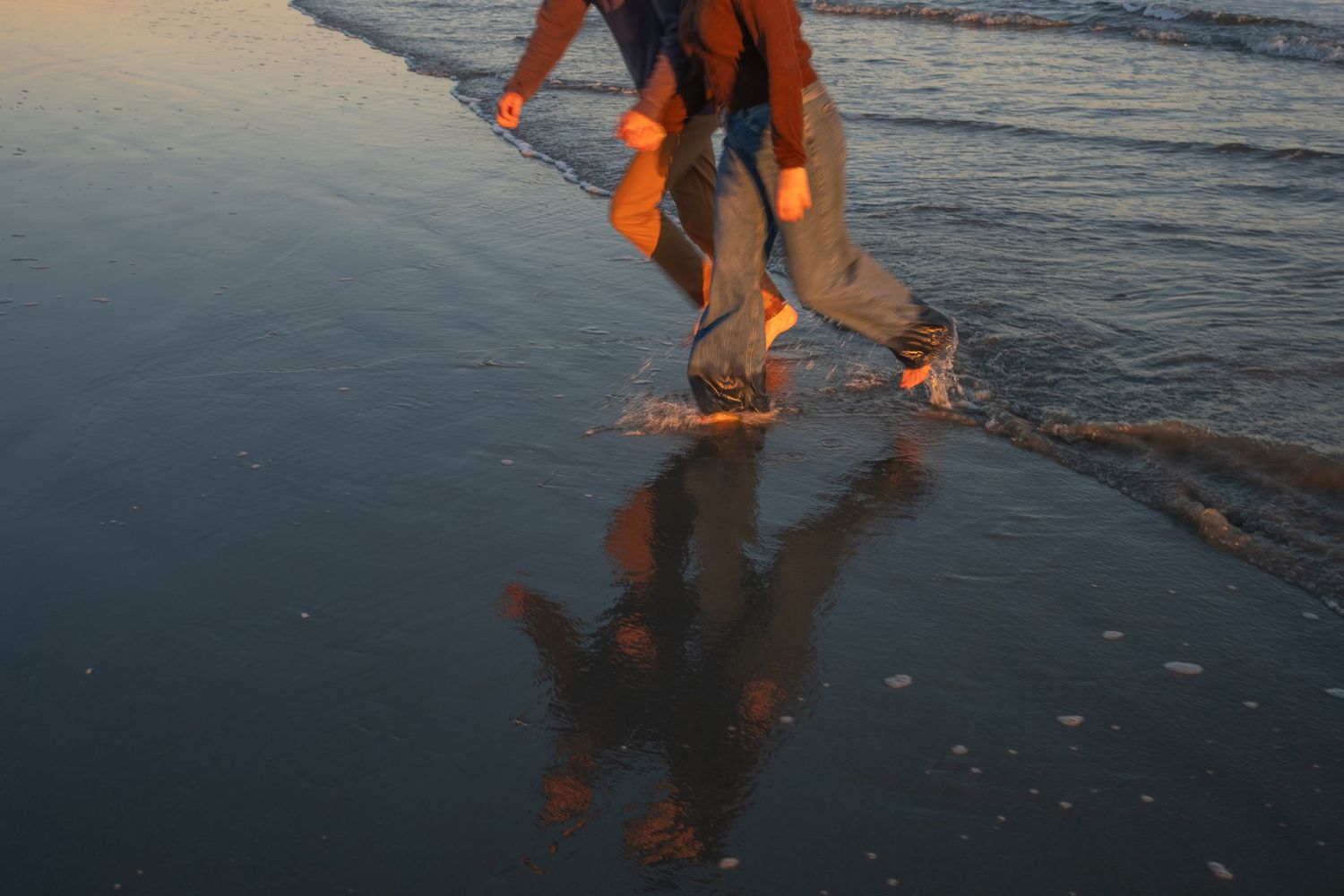 Barefoot person with rolled-up jeans walking through shallow ocean water at sunset, with a golden reflection on the wet sand.