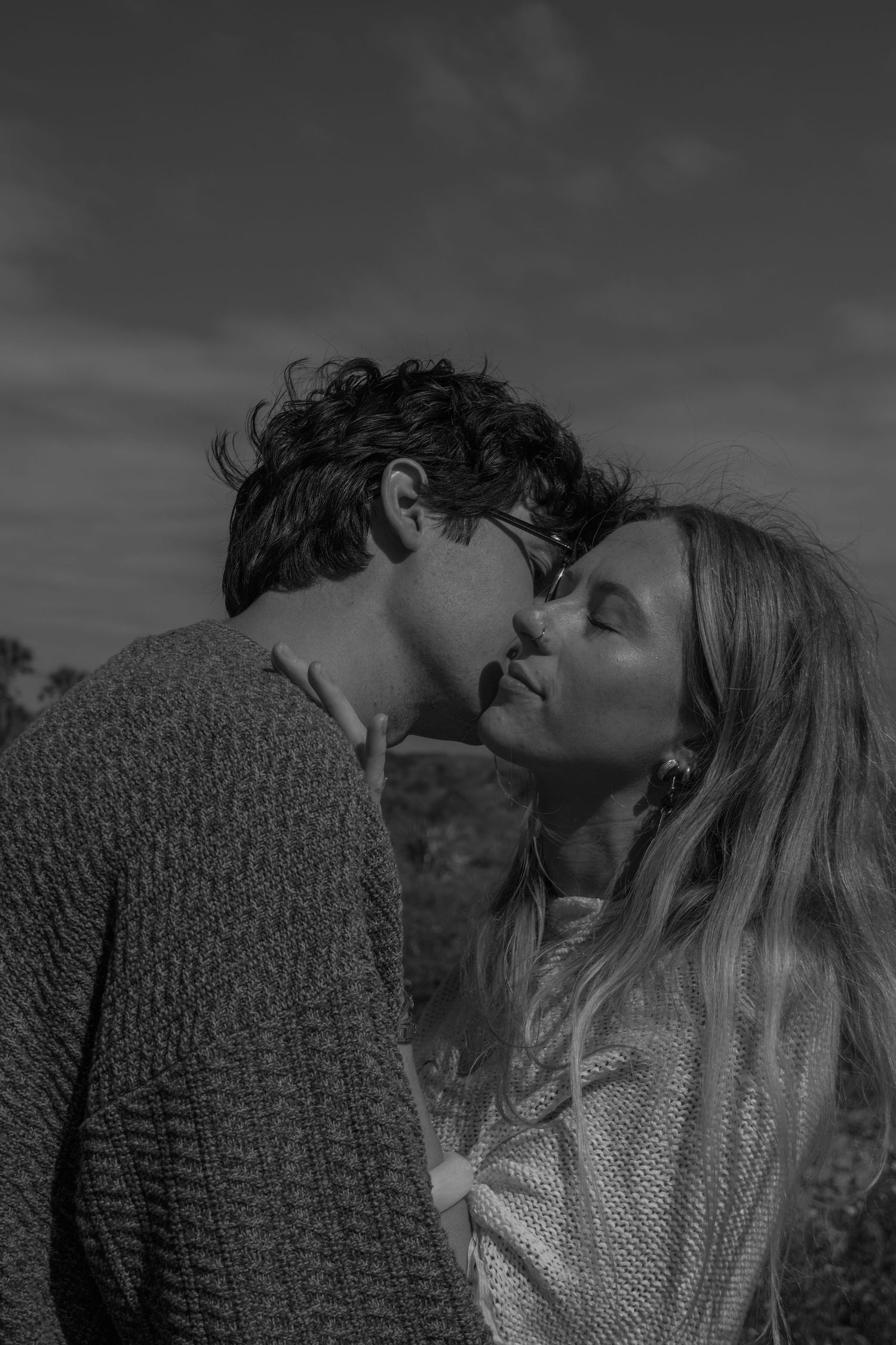 A black and white portrait of a young couple about to kiss, the man in a knit sweater and glasses, the woman with long wavy hair.