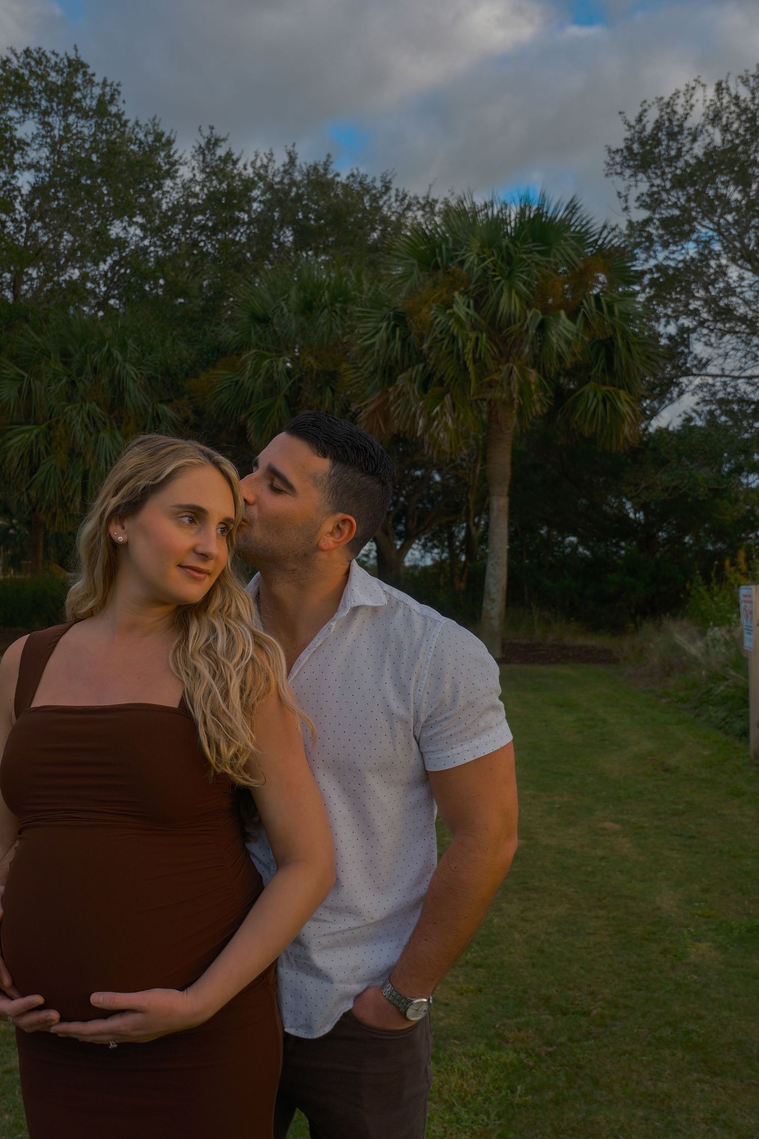 Man kisses the cheek of his pregnant partner in a brown dress as they pose outdoors near palm trees.