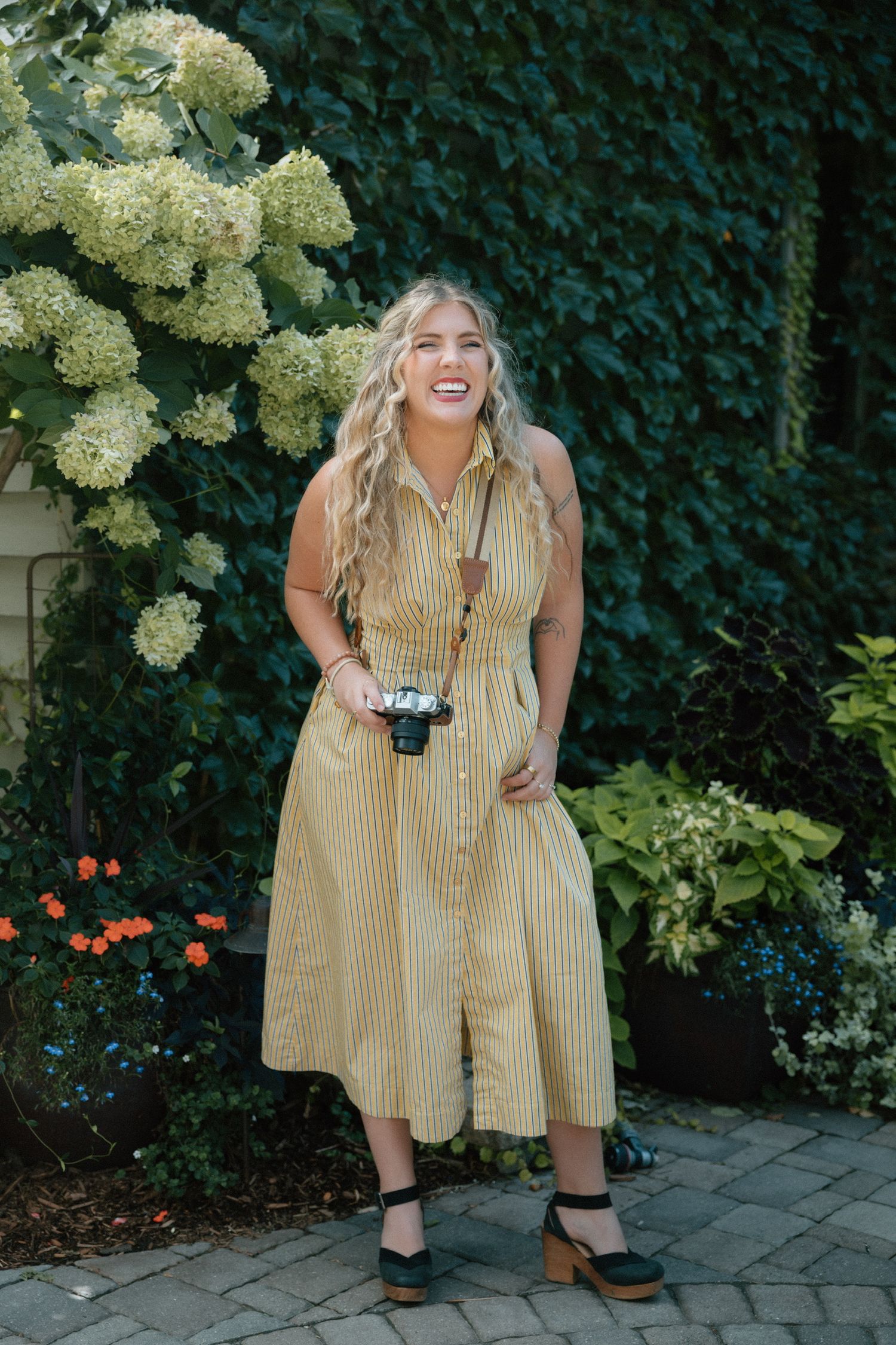 Laughing woman in a long yellow striped dress and platform sandals holds a film camera in a garden with hydrangeas and ivy.