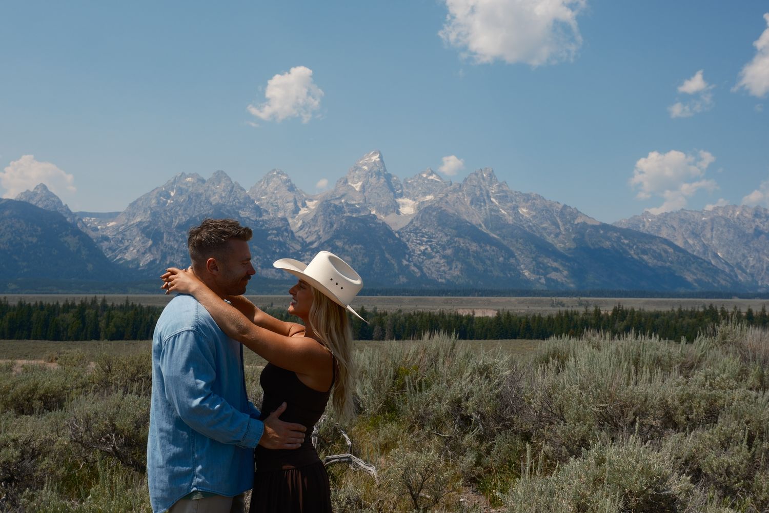 Couple embracing and smiling at each other in an open meadow with the snowy Teton mountain range behind them.
