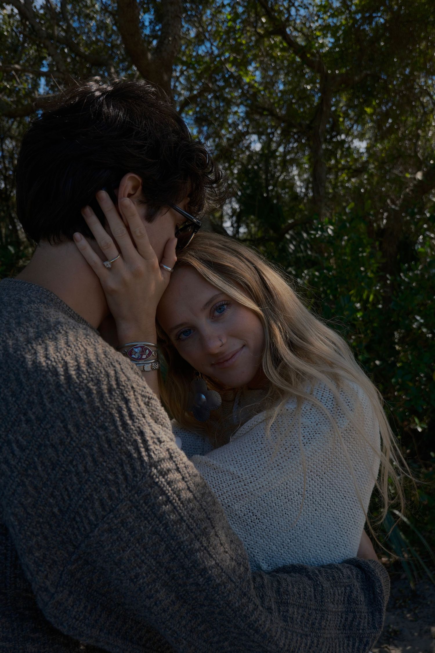 A man in a cozy knit sweater gently cradles a smiling blonde woman's head as they embrace beneath leafy trees.