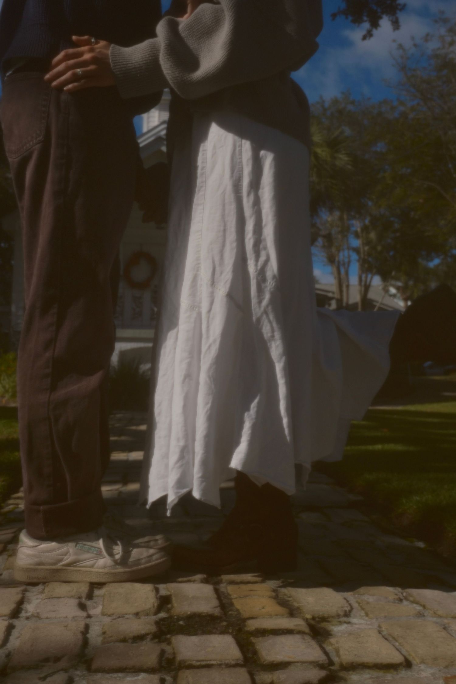 Close-up of a couple standing together on a brick path, her white flowy skirt and his brown trousers in sunlight.