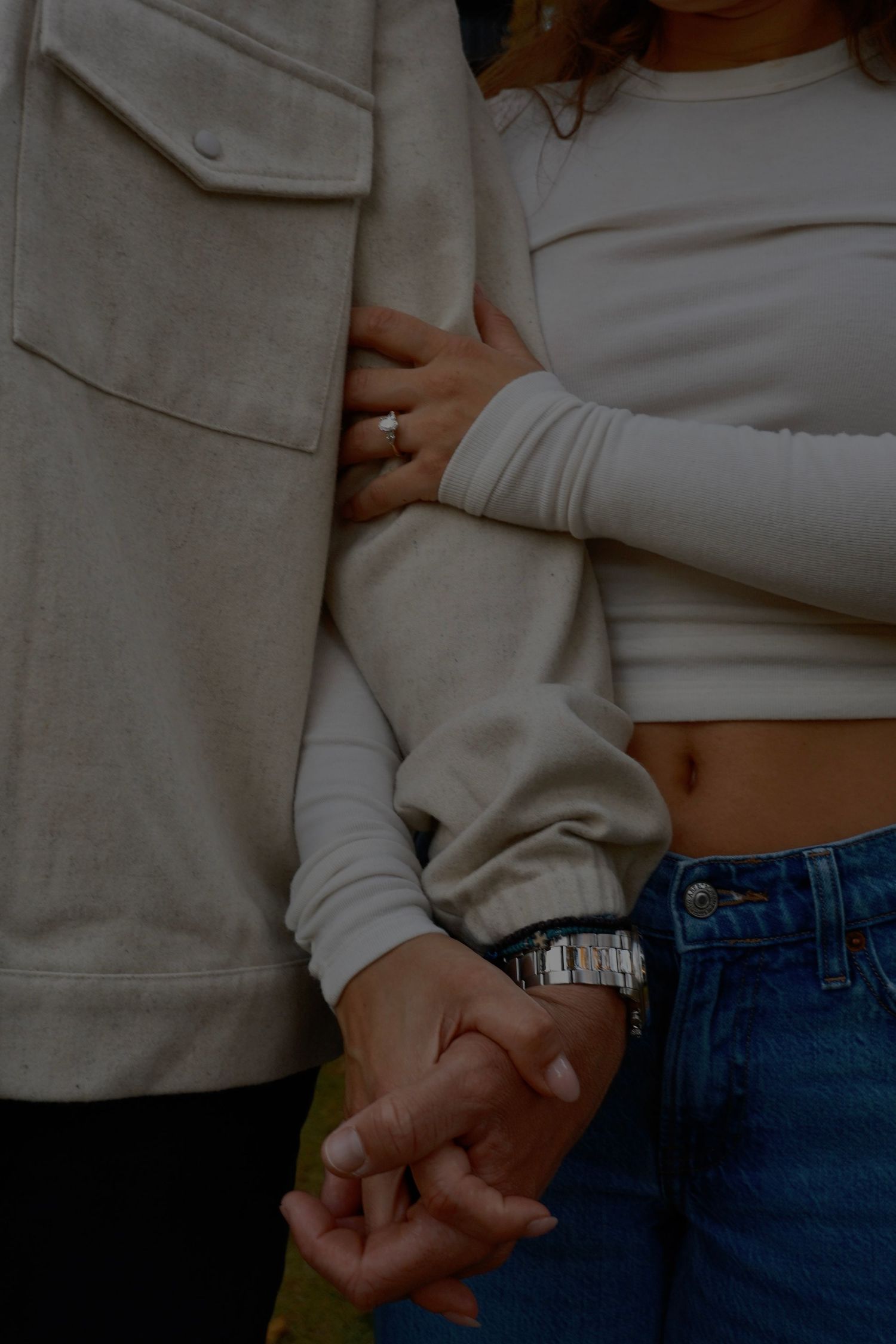 Close-up of a couple holding hands, the woman in a cream top and jeans wearing a ring while the man wears a beige jacket and watch.