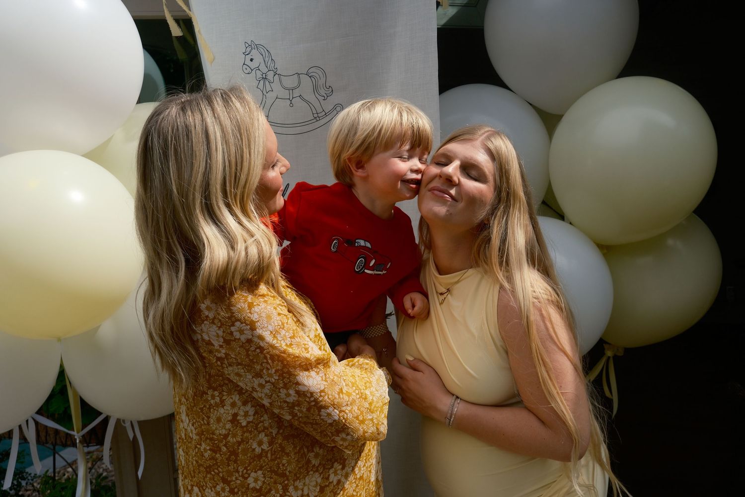 A woman kisses the cheek of a young boy in red while a pregnant woman smiles beside them among cream balloons.