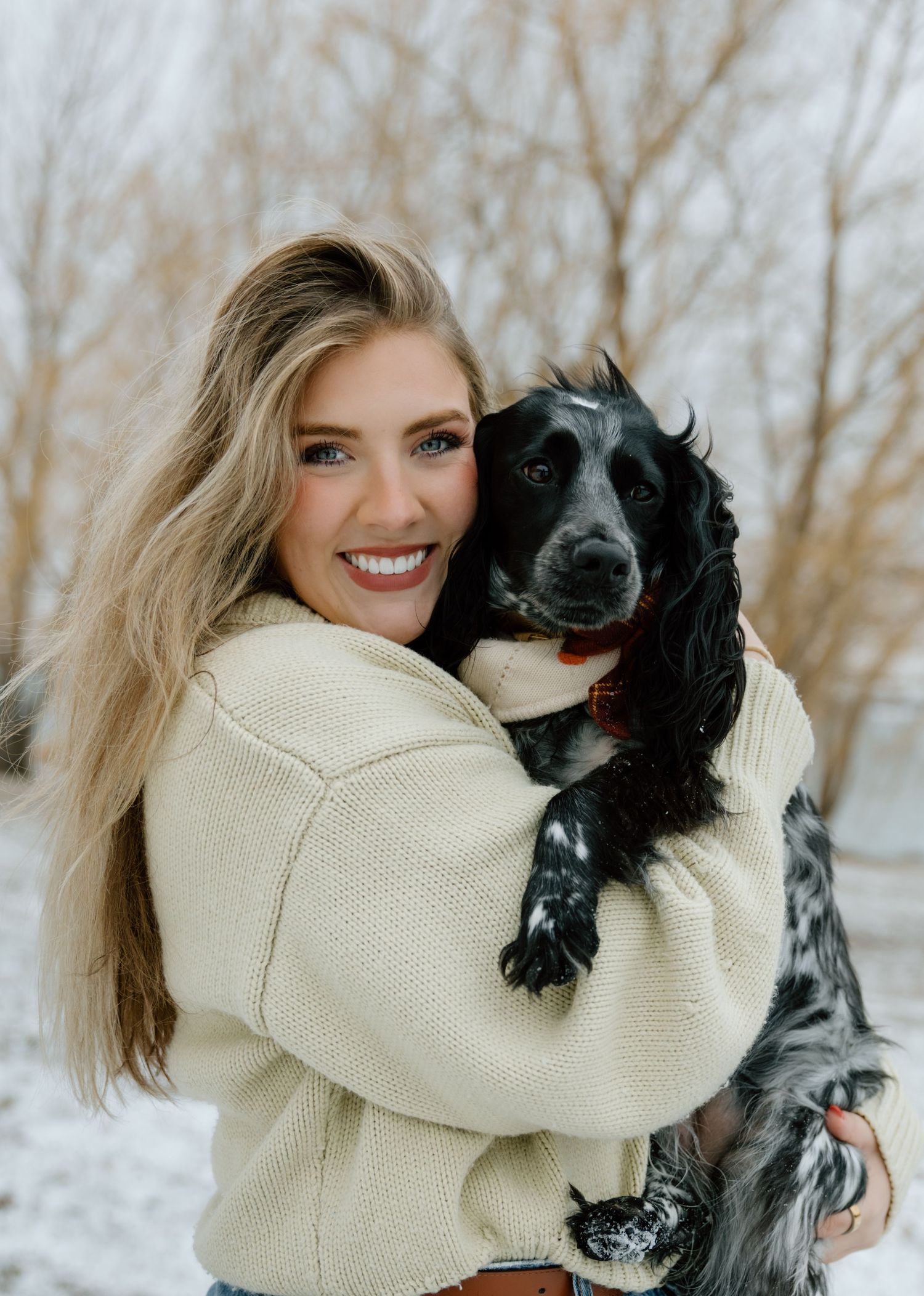 A smiling blonde woman in a cream knit sweater hugs a black and white spaniel in a snowy landscape with bare winter trees.