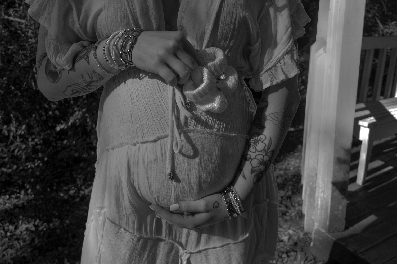 Black and white maternity photo of a pregnant woman in a flowing dress with bracelets and tattoos, cradling her belly.