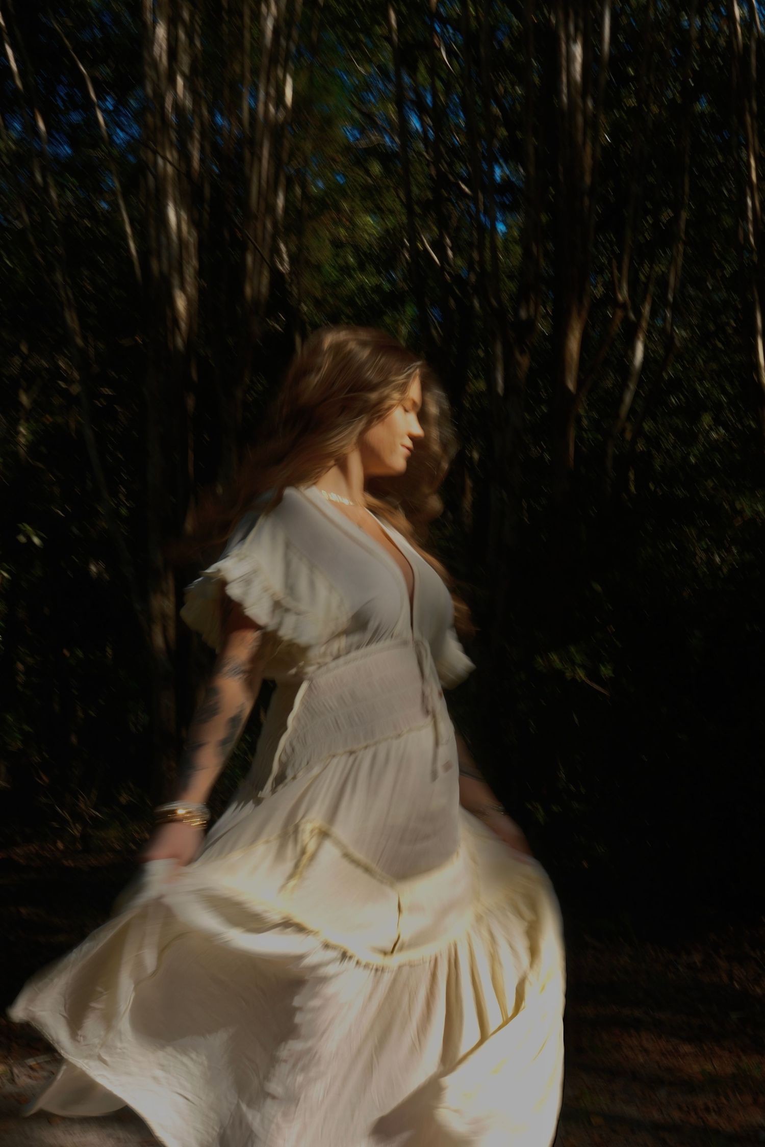 Woman in a flowing cream dress twirls in a dark forest, her hair and skirt caught in soft motion blur.