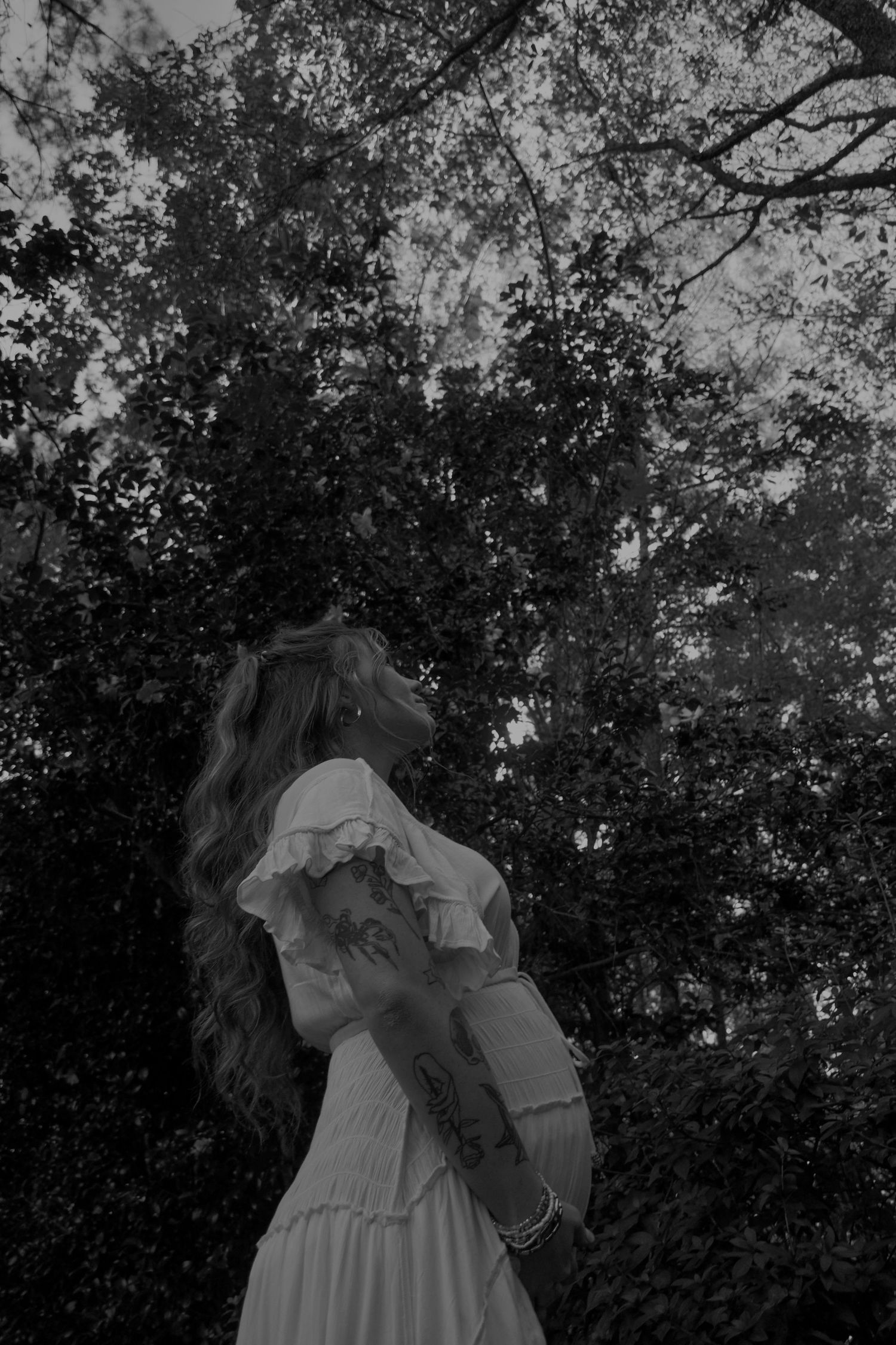 Black and white photo of a pregnant woman with long wavy hair and arm tattoos in a ruffled dress, gazing up at tall forest trees.