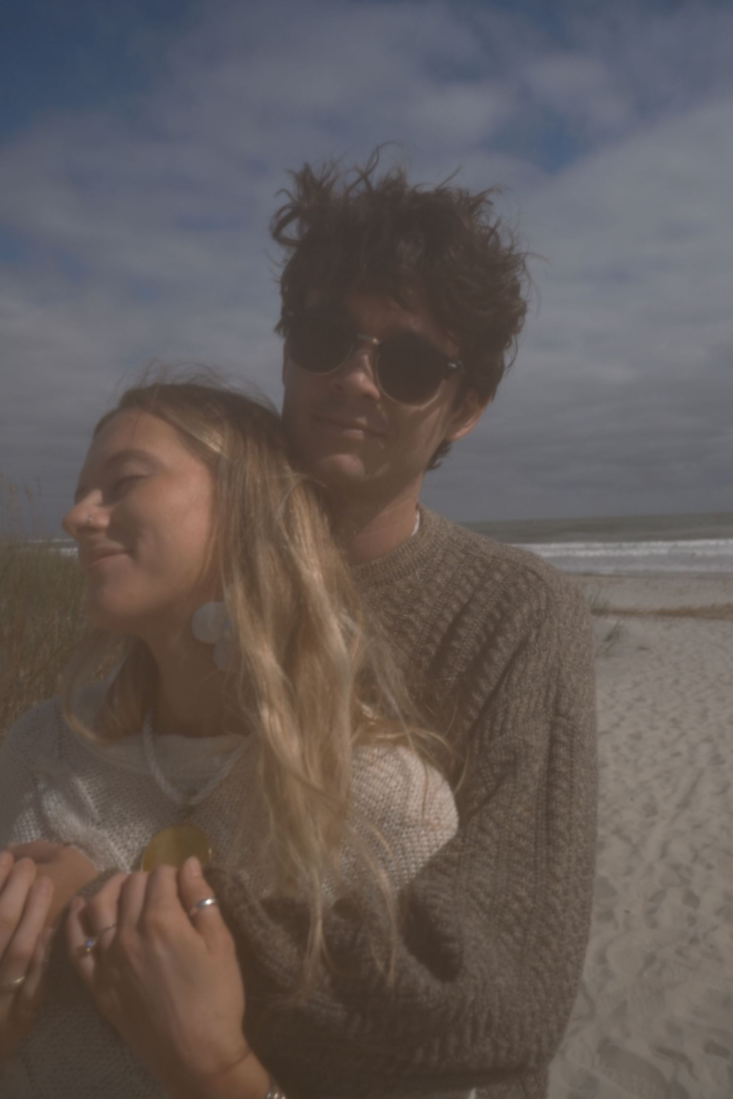 A couple embraces on a windswept beach, the man in sunglasses and a knit sweater hugging a smiling woman with long blonde hair.