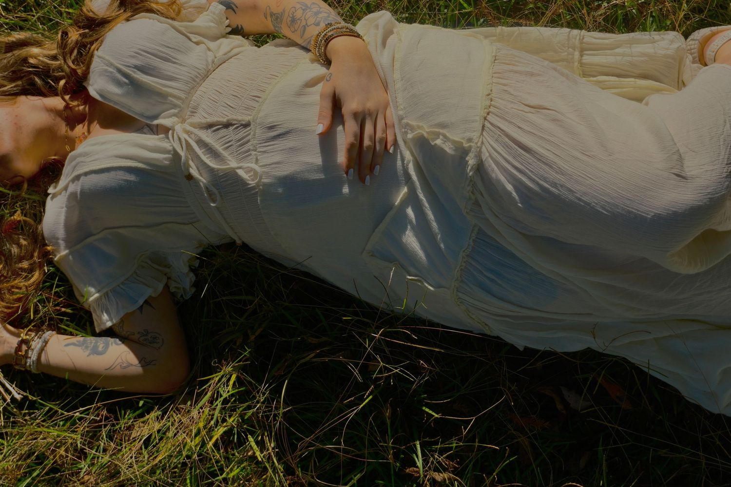 A pregnant woman in a white dress lies on sunlit grass with her hand resting on her belly in a maternity photo.