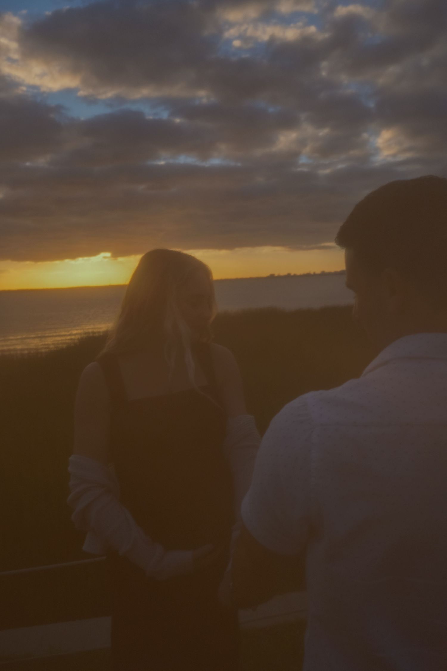 Silhouetted couple stands facing a colorful sunset over the water, with dramatic clouds in the sky.
