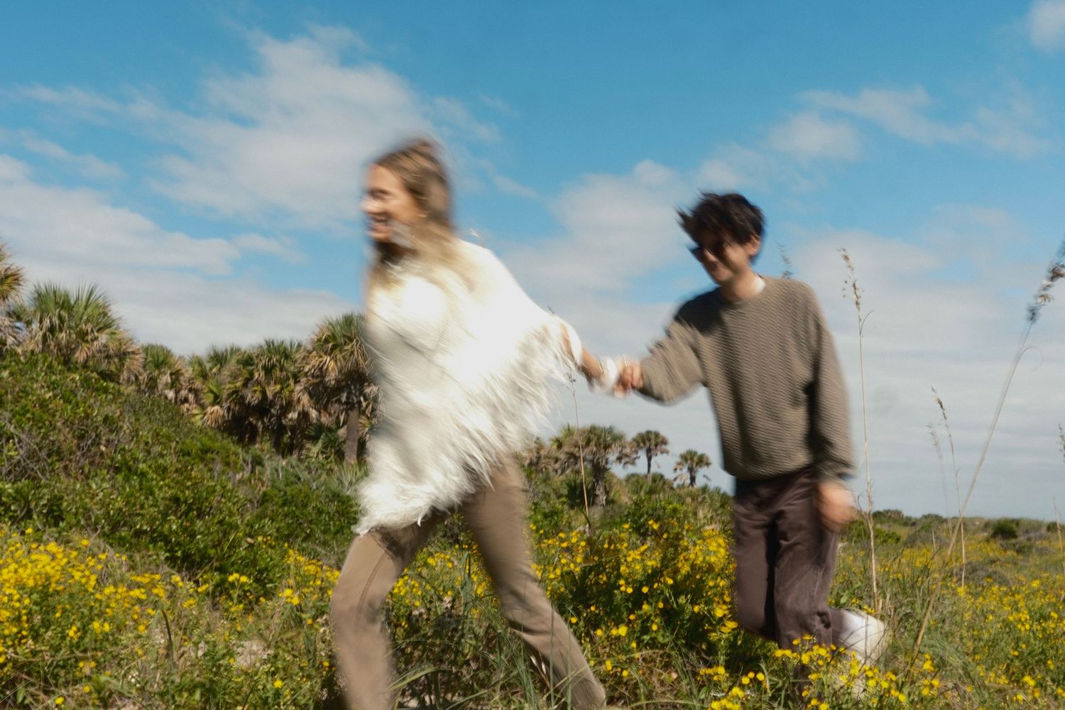 A smiling couple holds hands and walks through a field of yellow wildflowers and palm trees under a bright blue sky.