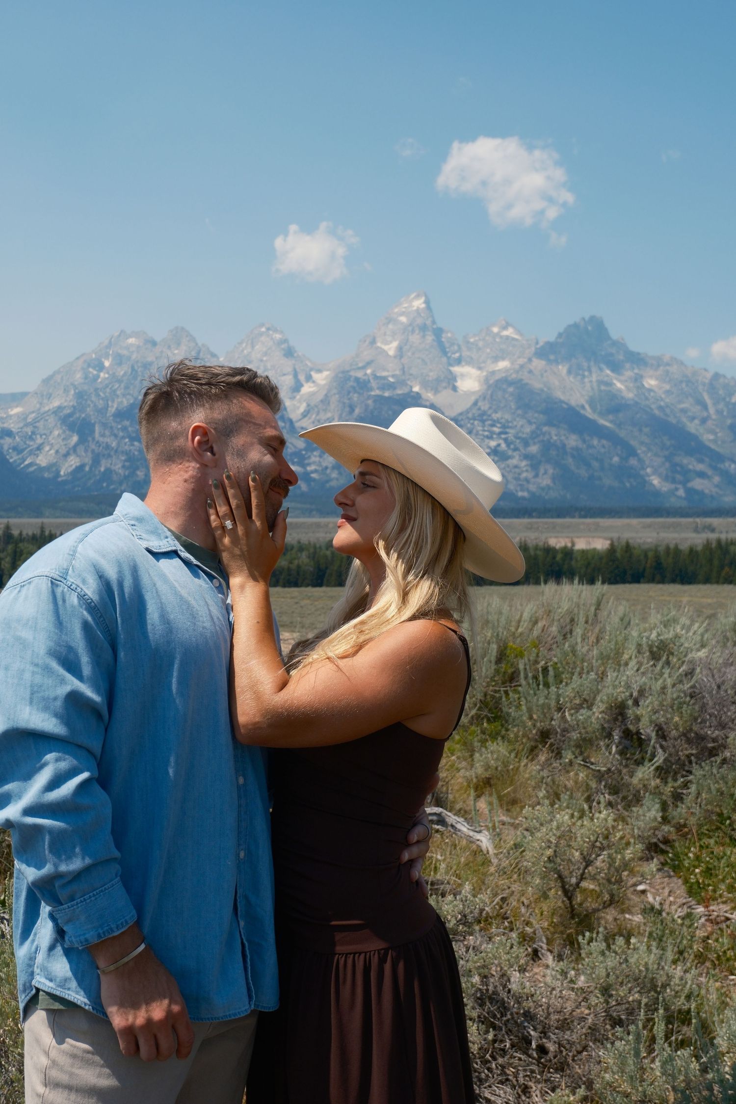 Woman in a white cowboy hat gently touches her partner's face as they share a romantic moment before the Teton mountains.
