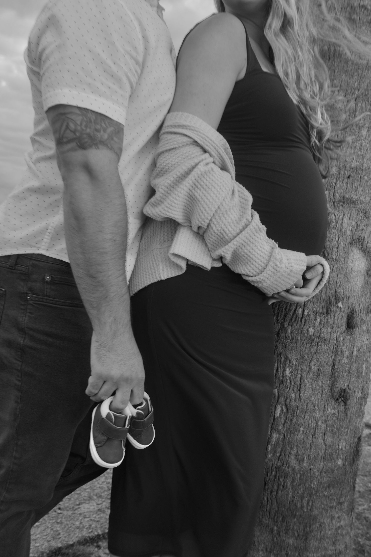 Black and white maternity portrait of a couple leaning on a tree, the man holding tiny baby shoes beside her baby bump.