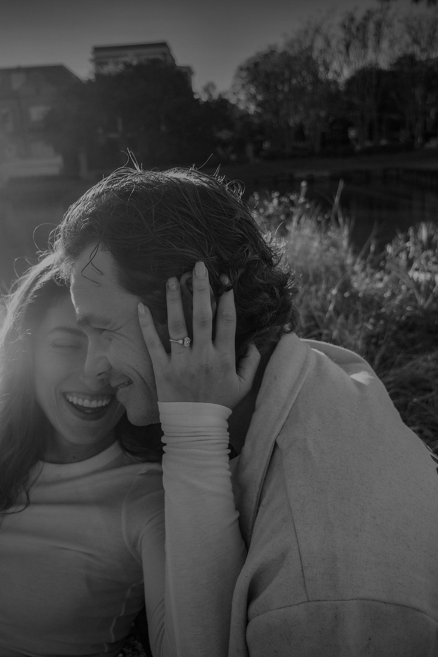 Black and white close-up of a laughing couple embracing outdoors, her hand with an engagement ring resting on his head.