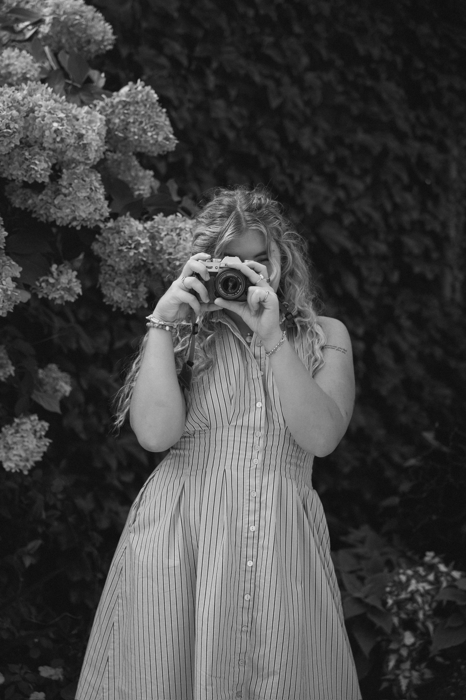 Black and white portrait of a woman in a striped button-front dress photographing with a film camera beside hydrangeas.