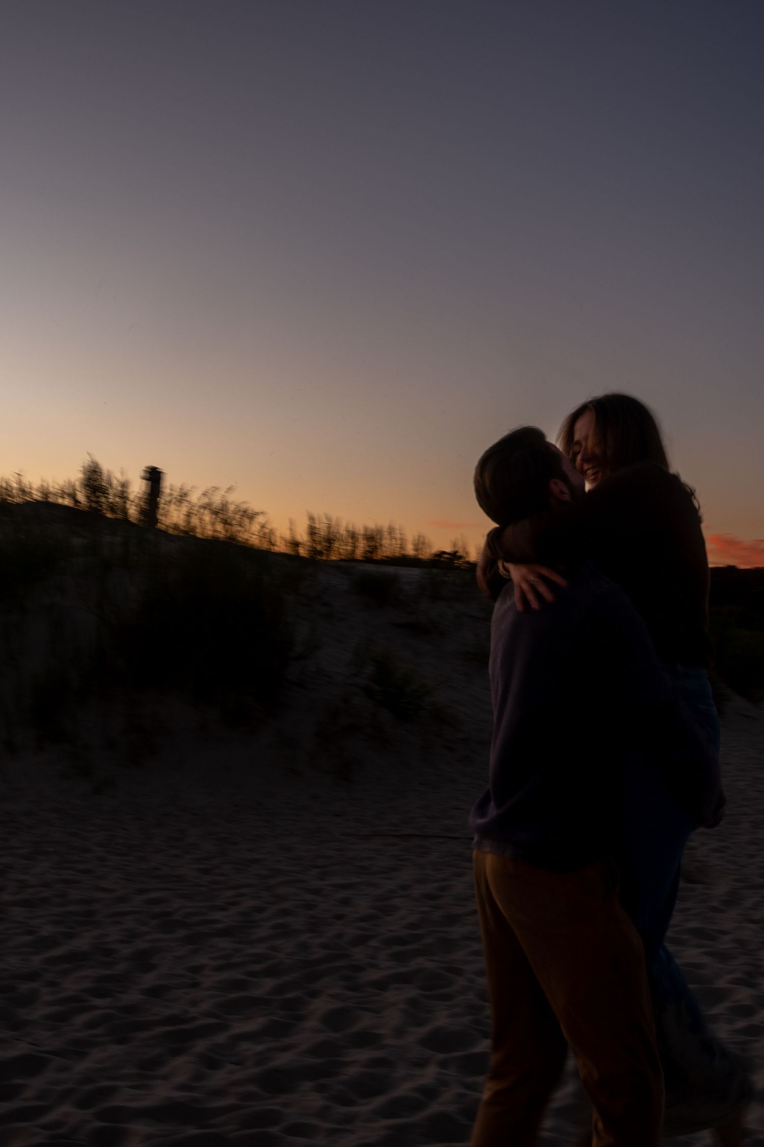Silhouetted couple embracing and kissing on a sandy beach dune at dusk, with a pink and purple sunset sky behind them.