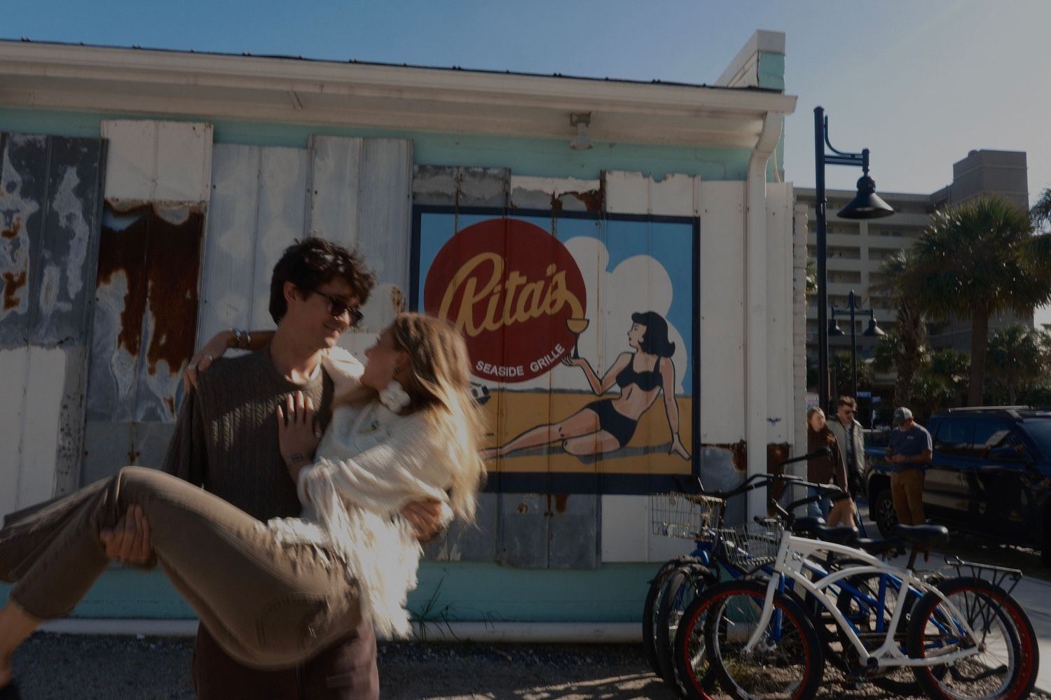 A man in sunglasses lifts a woman in a white top in a romantic embrace outside a beachside shop with a retro pin-up mural.