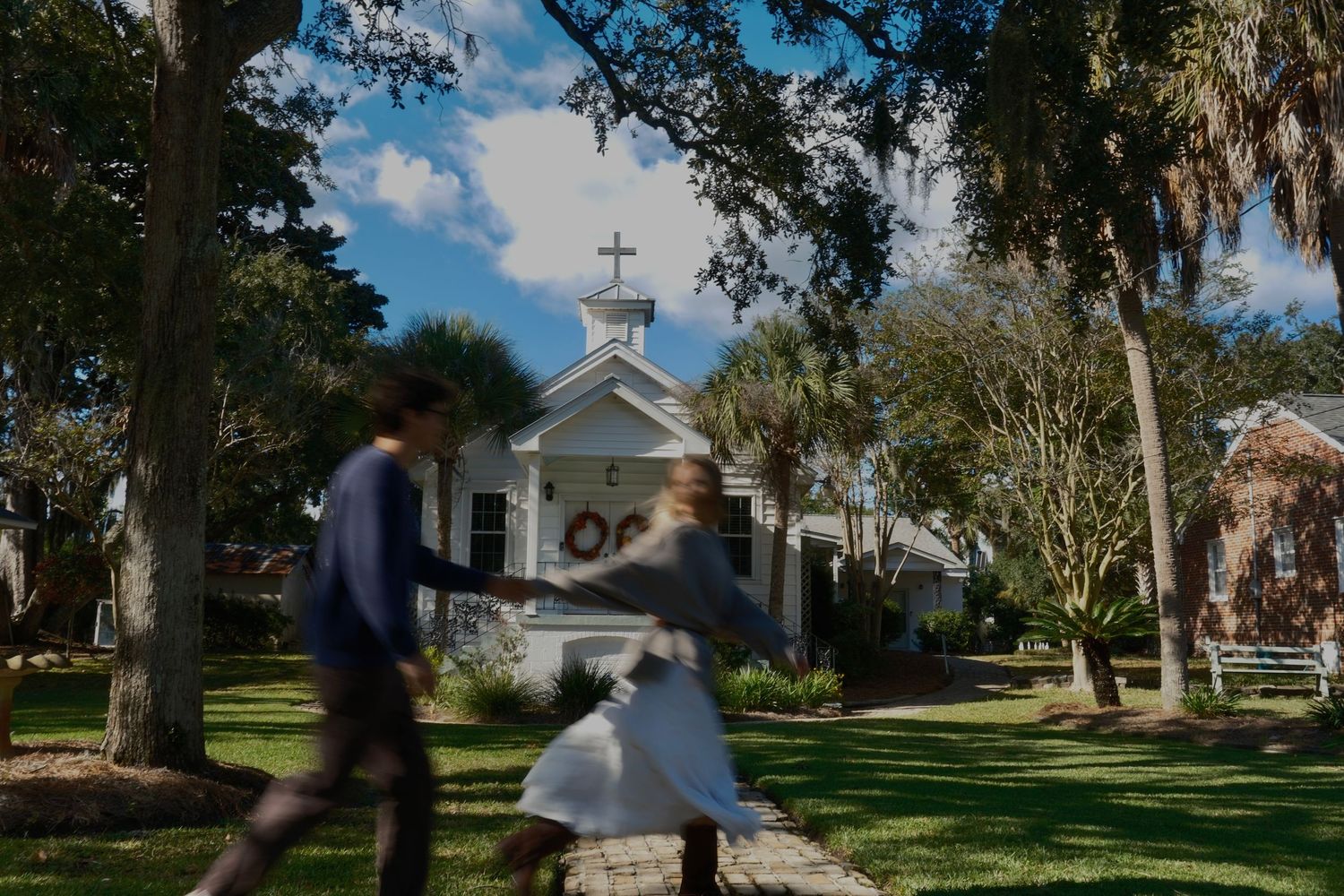 A couple holds hands and walks across a sunny green lawn toward a small white chapel framed by oaks and palm trees.