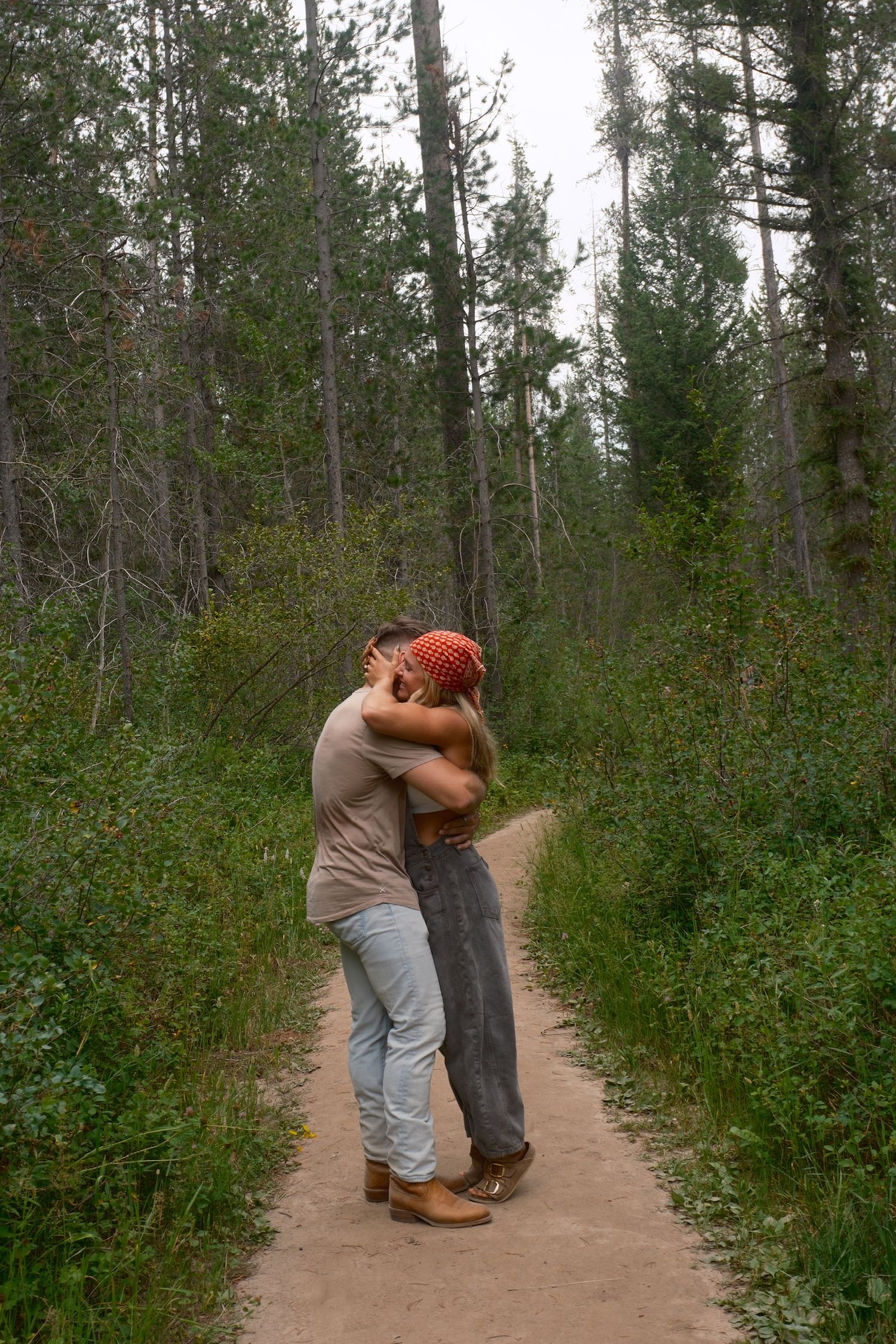 A couple embraces on a dirt path surrounded by tall pine trees and green forest underbrush.