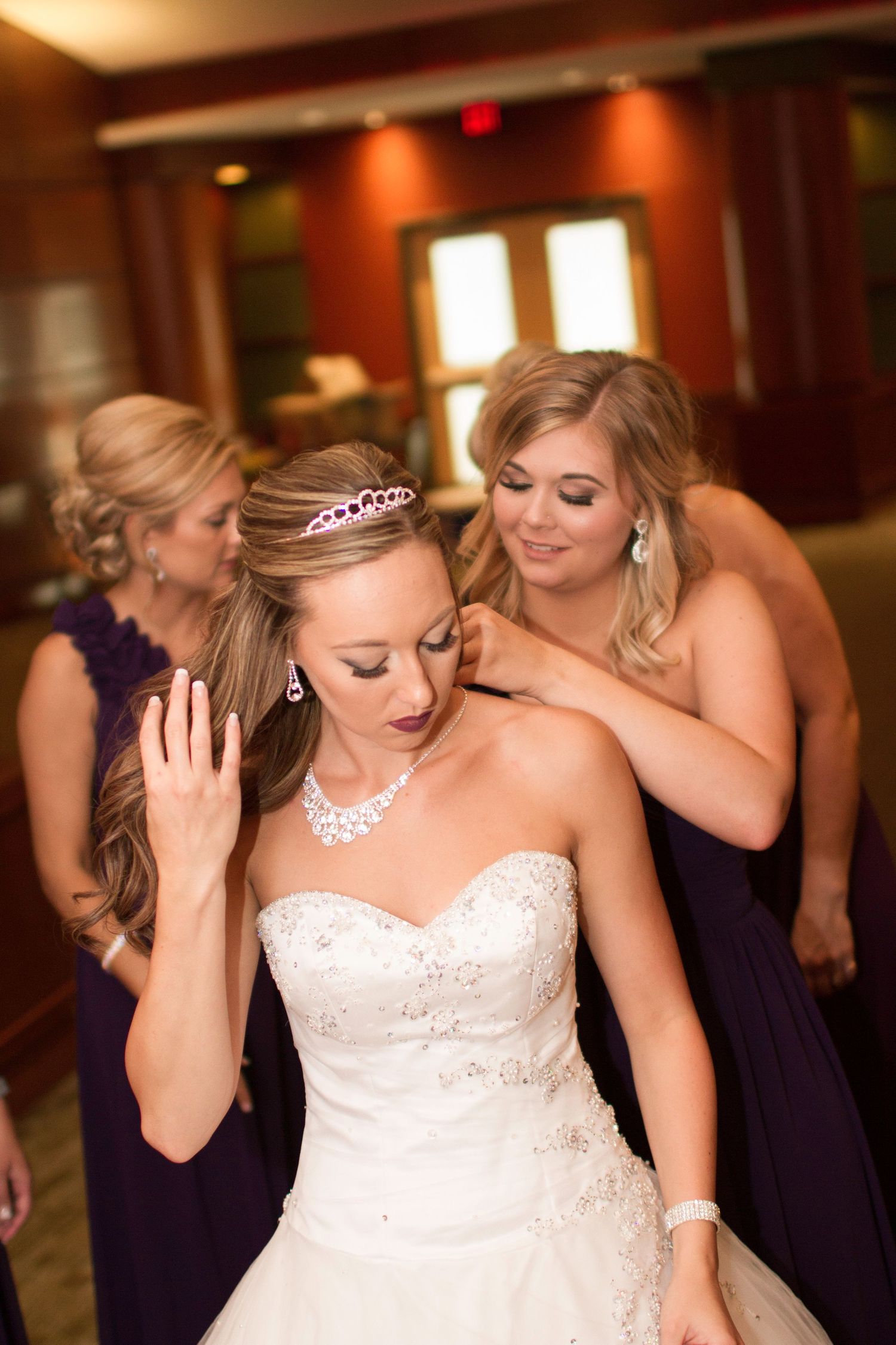 A bride in a strapless wedding gown and tiara gets ready with bridesmaids in purple dresses.