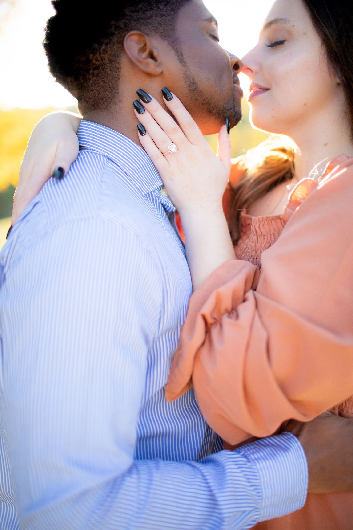 An intimate couple embraces in warm sunlight wearing blue striped shirt and peach colored top.