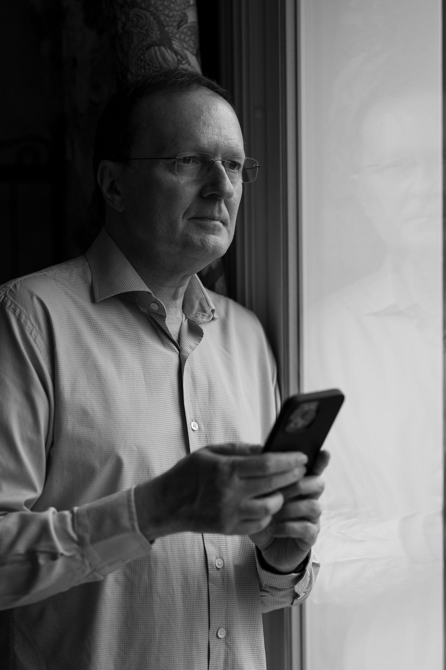 Black and white portrait of photographer Philippe Triconnet holding a phone beside a window.