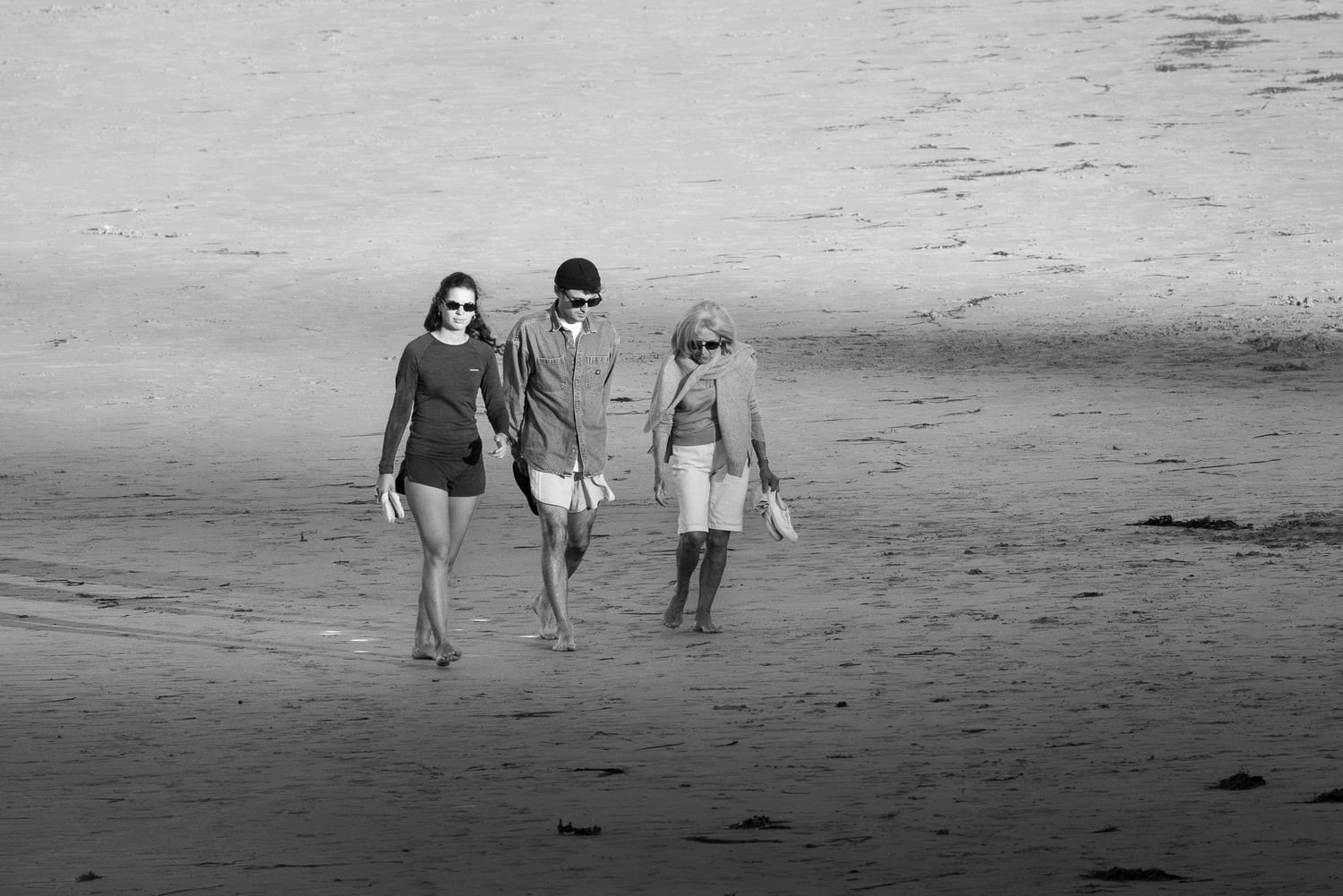 Un couple et une belle-maman marchent pieds nus sur la plage des Sables Blancs à Tréboul, à Douarnenez. Photographie en noir et blanc.