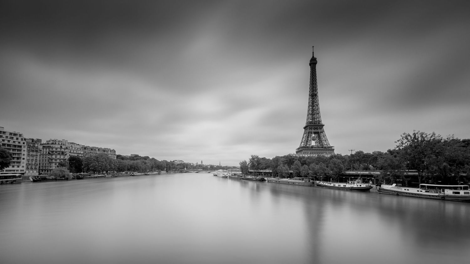 Black and white view of the Eiffel Tower and the Seine beneath a cloudy sky in Paris.