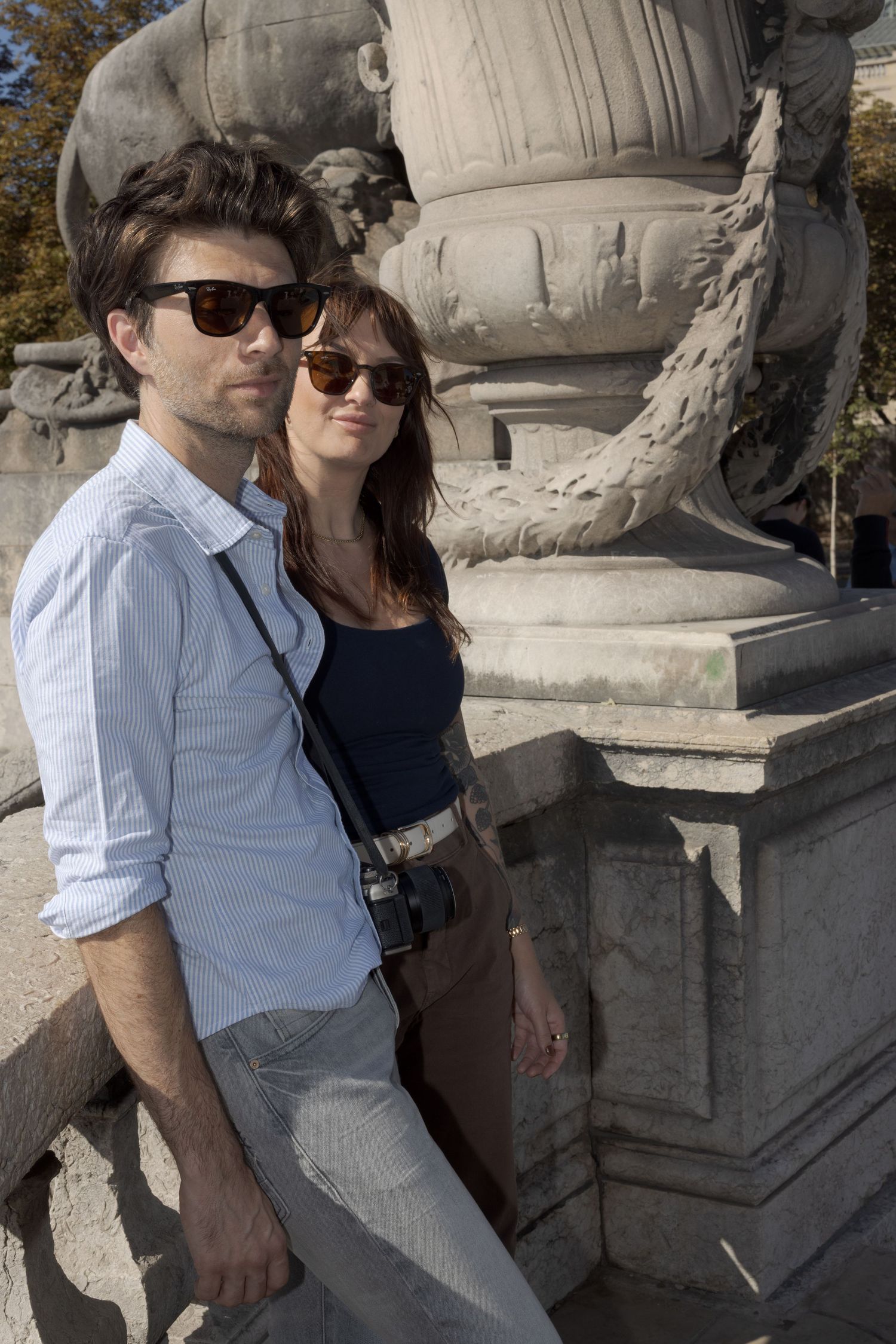 A couple wearing sunglasses posing beside a carved stone sculpture in Paris, Pont Alexandre III.
